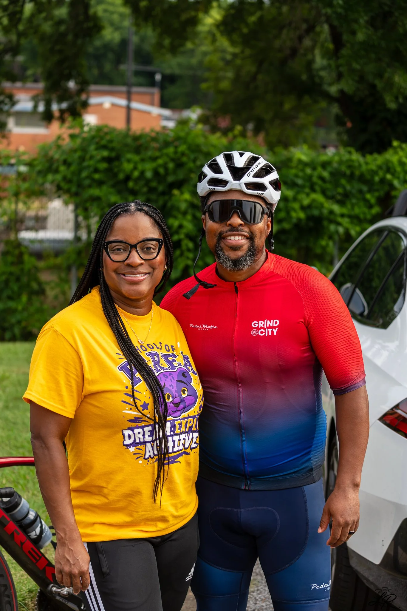 Two smiling people, a woman with glasses wearing a yellow T-shirt and a man with a beard and sunglasses wearing a red cycling jersey and helmet, posing outdoors near a vehicle and bicycle.