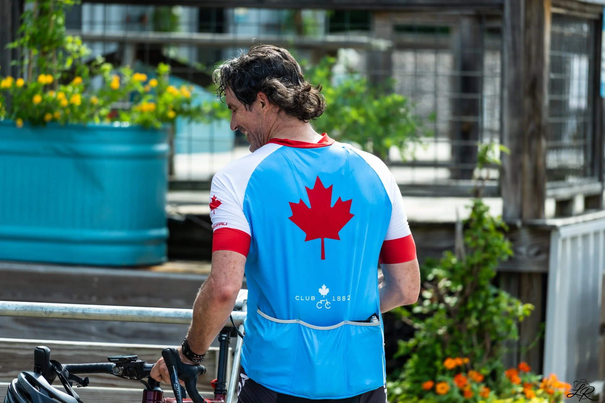 A man wearing a sports jersey with a red maple leaf on the back walking his bike outdoors in a garden with flowers.