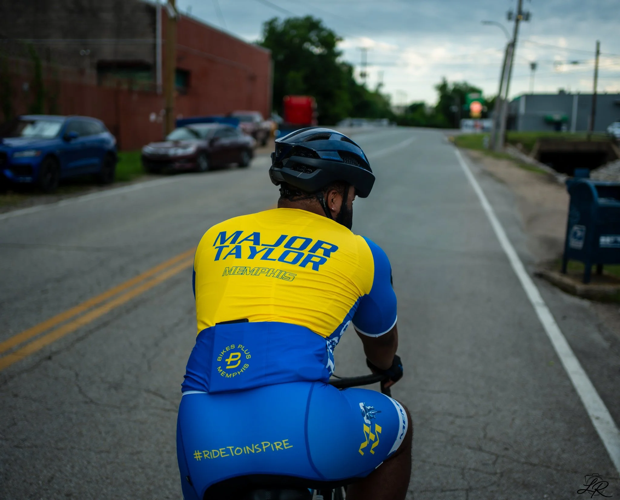 A cyclist wearing a blue and yellow jersey with the hashtag #RIDETOINSPIRE and 'Memphis' written on it, riding on a street during daytime with parked cars, utility poles, and buildings in the background.