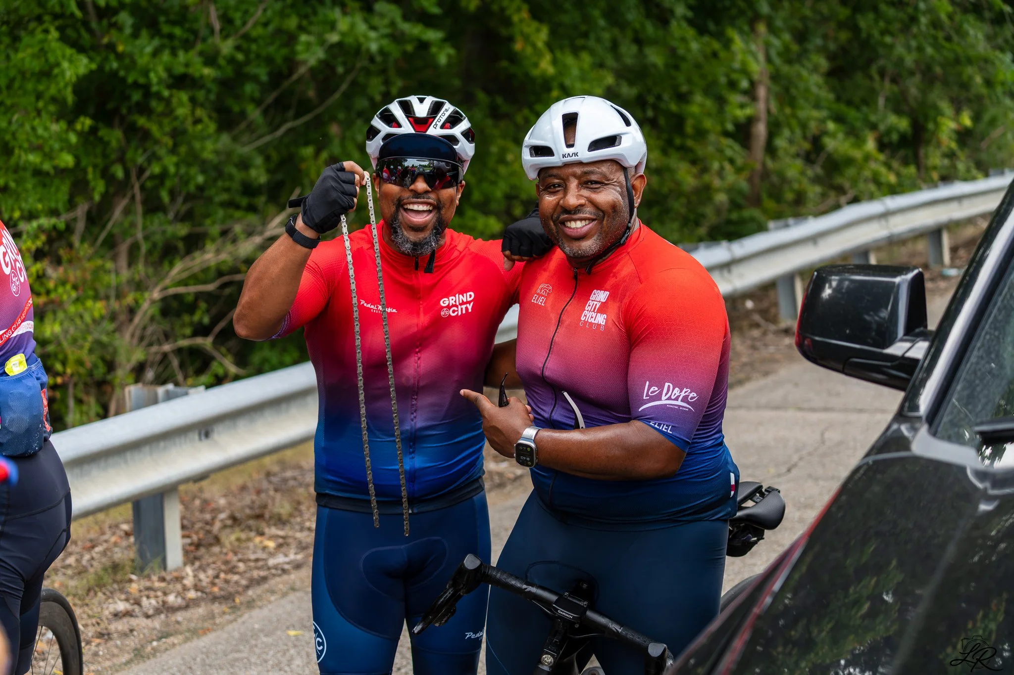 Two smiling men wearing cycling helmets and red and purple cycling jerseys, standing next to a black vehicle and a bicycle on a roadside with green trees in the background.