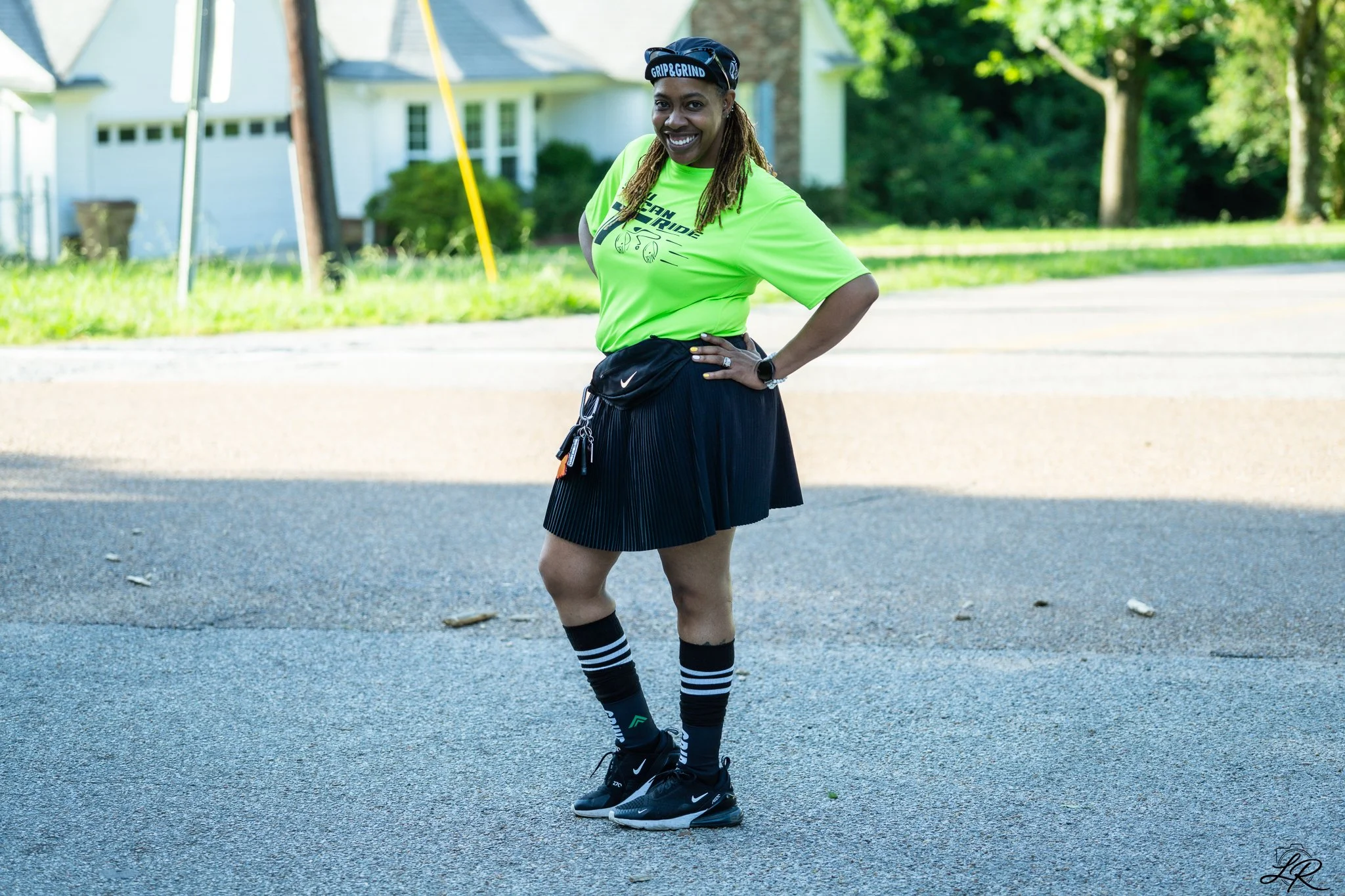 A woman standing on a street during daytime, smiling, dressed in athletic gear including a neon green T-shirt, a black pleated skirt, striped knee-high socks, and black sneakers, with a black fanny pack around her waist, and a black headband with whi