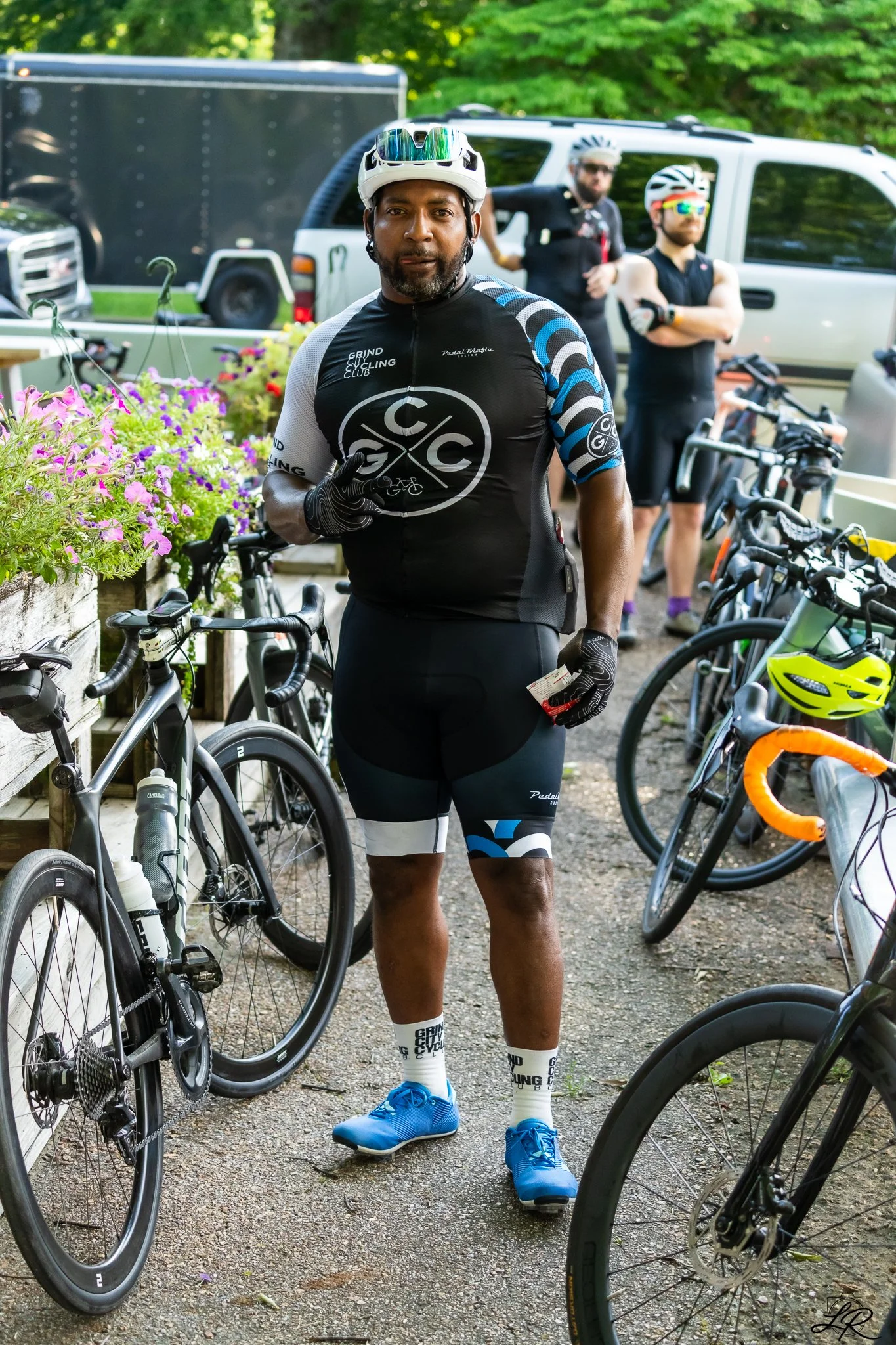 A man in cycling gear standing among parked bicycles, with two other cyclists in the background, outdoors surrounded by cars, trucks, and greenery.