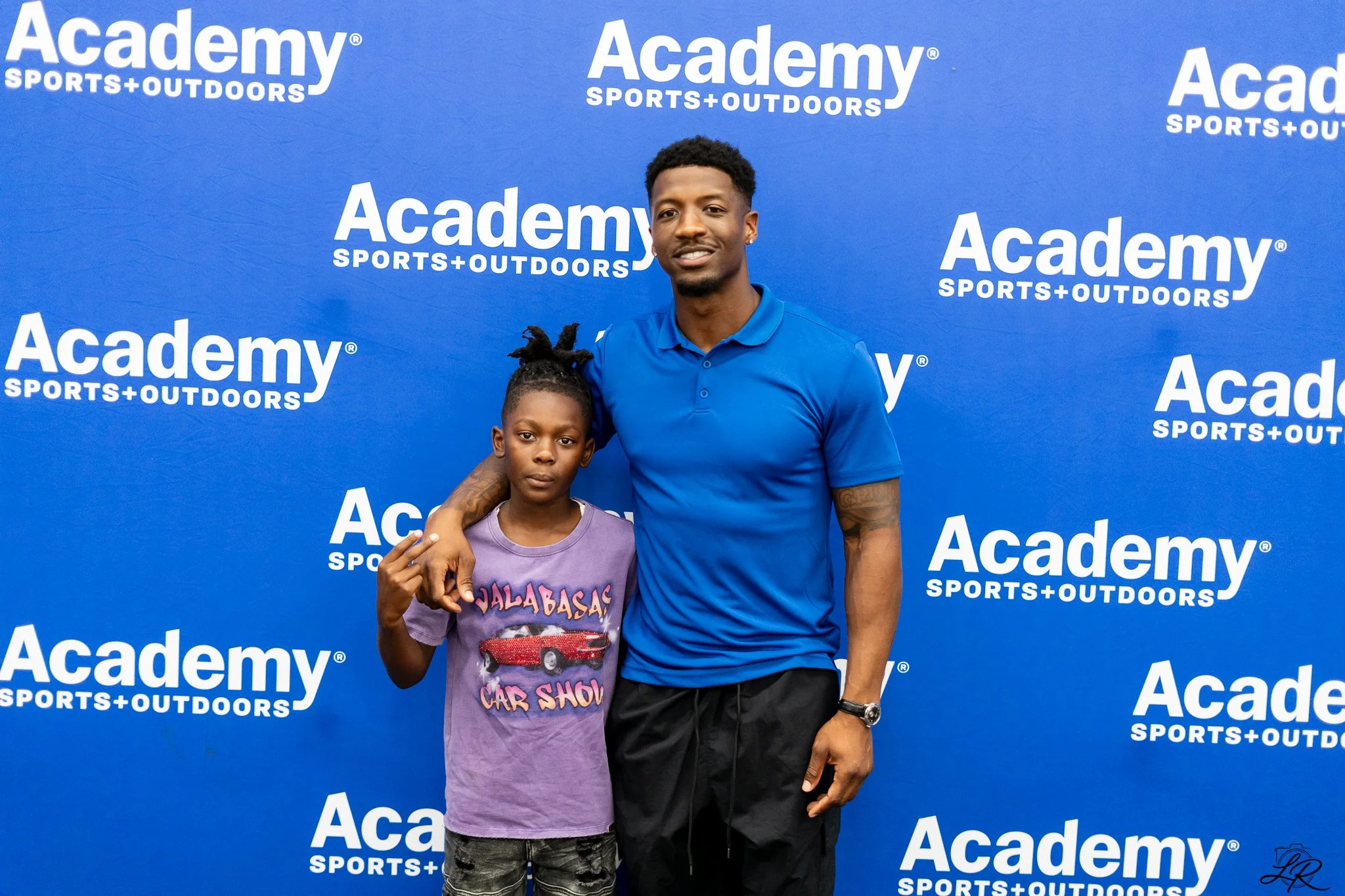 A young man and a young girl standing in front of a blue backdrop with White text that says 'Academy Sports + Outdoors'. The young man is wearing a blue polo shirt and black pants with a wristwatch, and the girl is wearing a purple T-shirt with graph