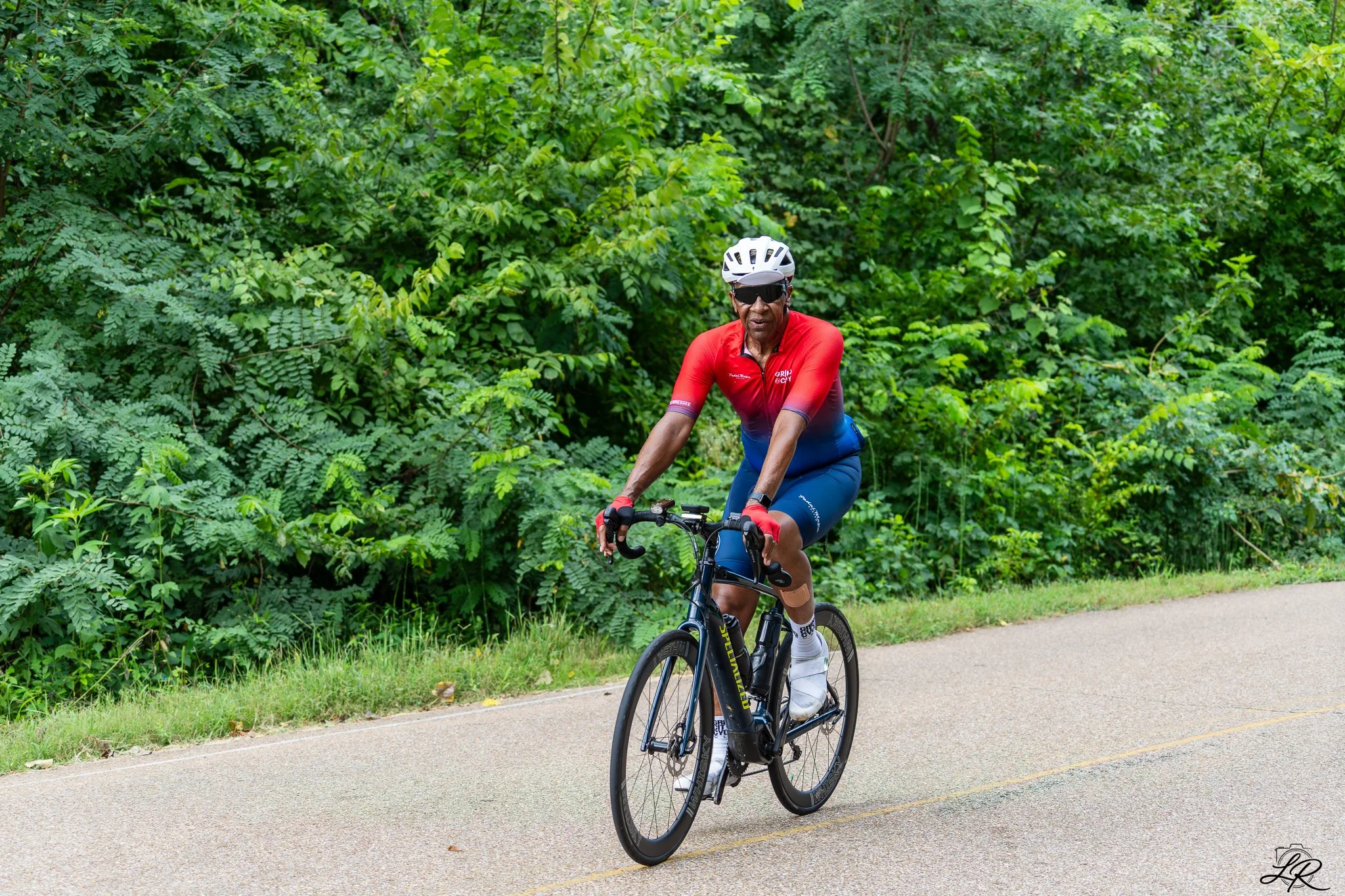 An elderly man in a red cycling shirt, black helmet, and sunglasses riding a black bicycle on a paved path surrounded by lush green foliage.