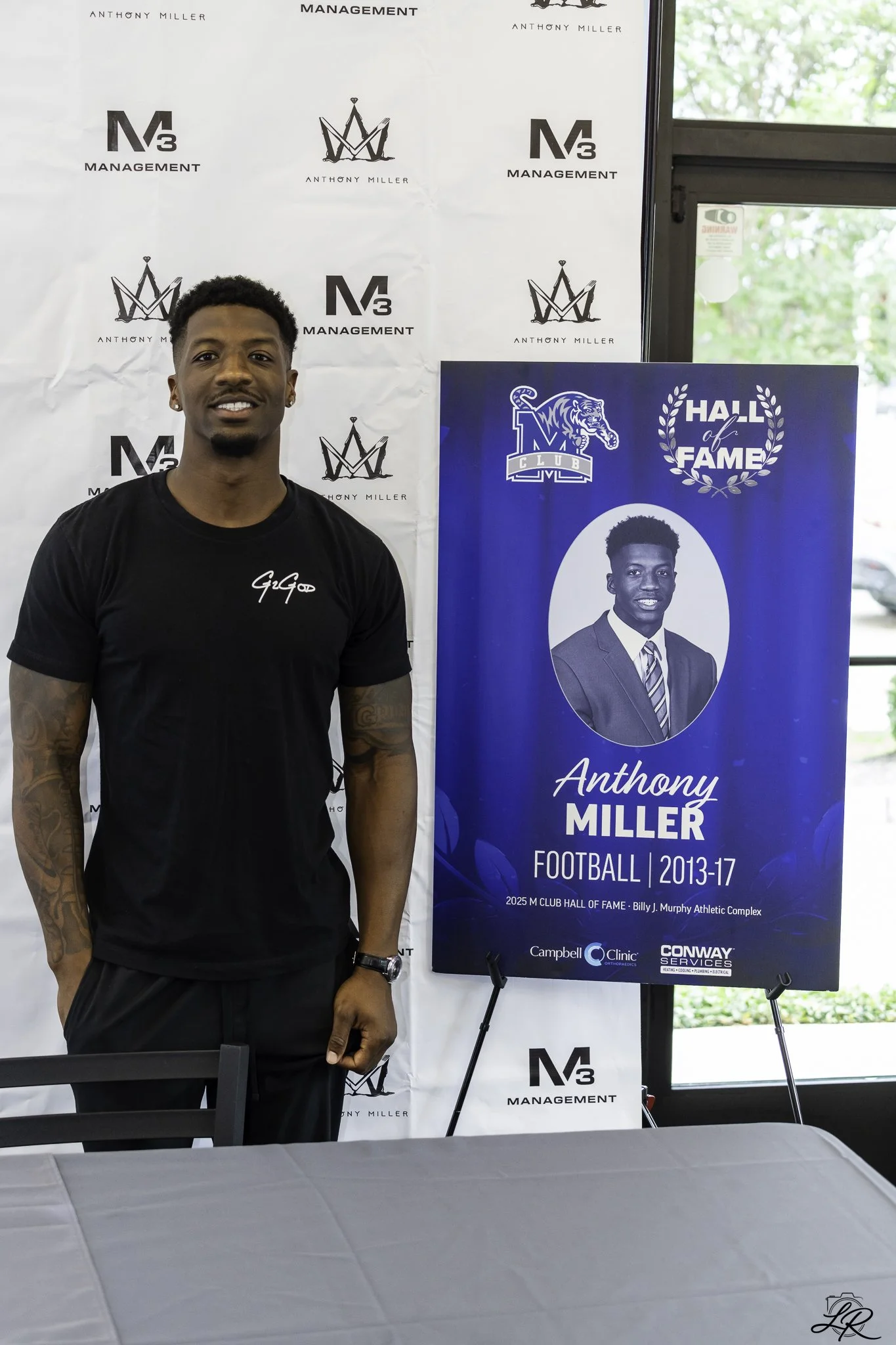 A young man with short curly hair, earrings, and a black t-shirt standing next to a blue poster. The poster features a black and white photograph of him, his name Anthony Miller, and text indicating he played football from 2013 to 2017. He is smiling