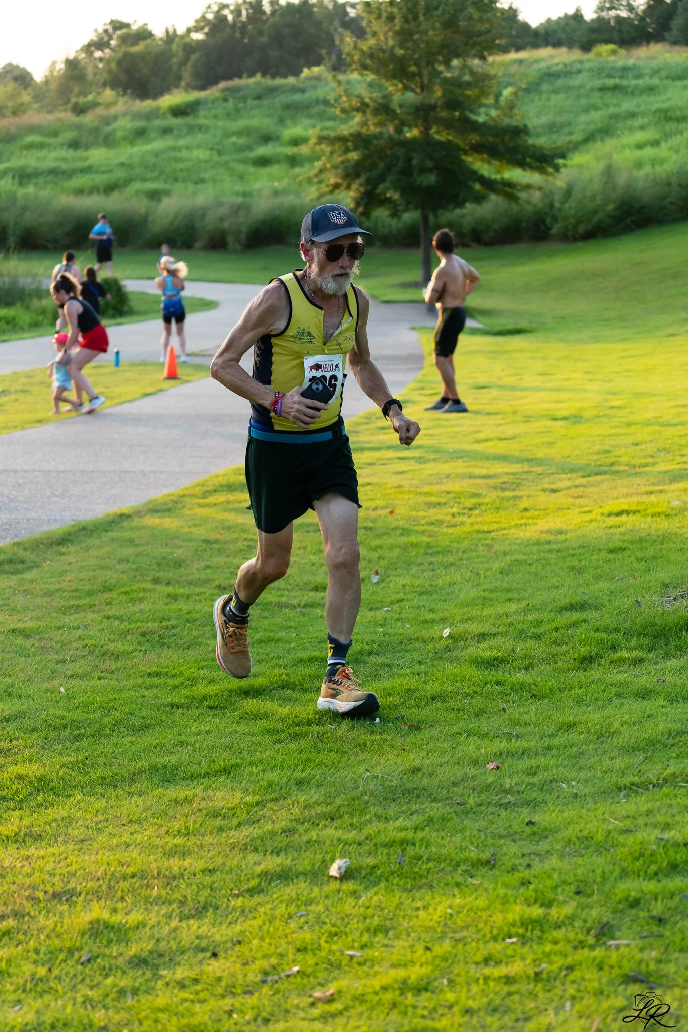 An elderly man running on a grassy area during a race, wearing a yellow and black running shirt, shorts, sunglasses, a cap, and holding a phone, with other runners and spectators in the background, lush green landscape, and sunset lighting.