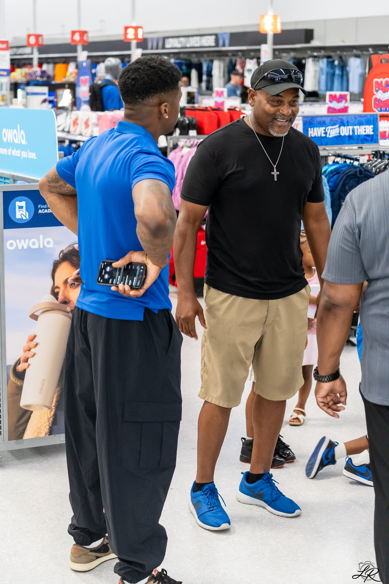 Two men talk inside a retail store, with clothing and signs in the background. One man is wearing a black t-shirt, khaki shorts, and a black cap with sunglasses, while the other is in a blue shirt and black pants, holding a smartphone.