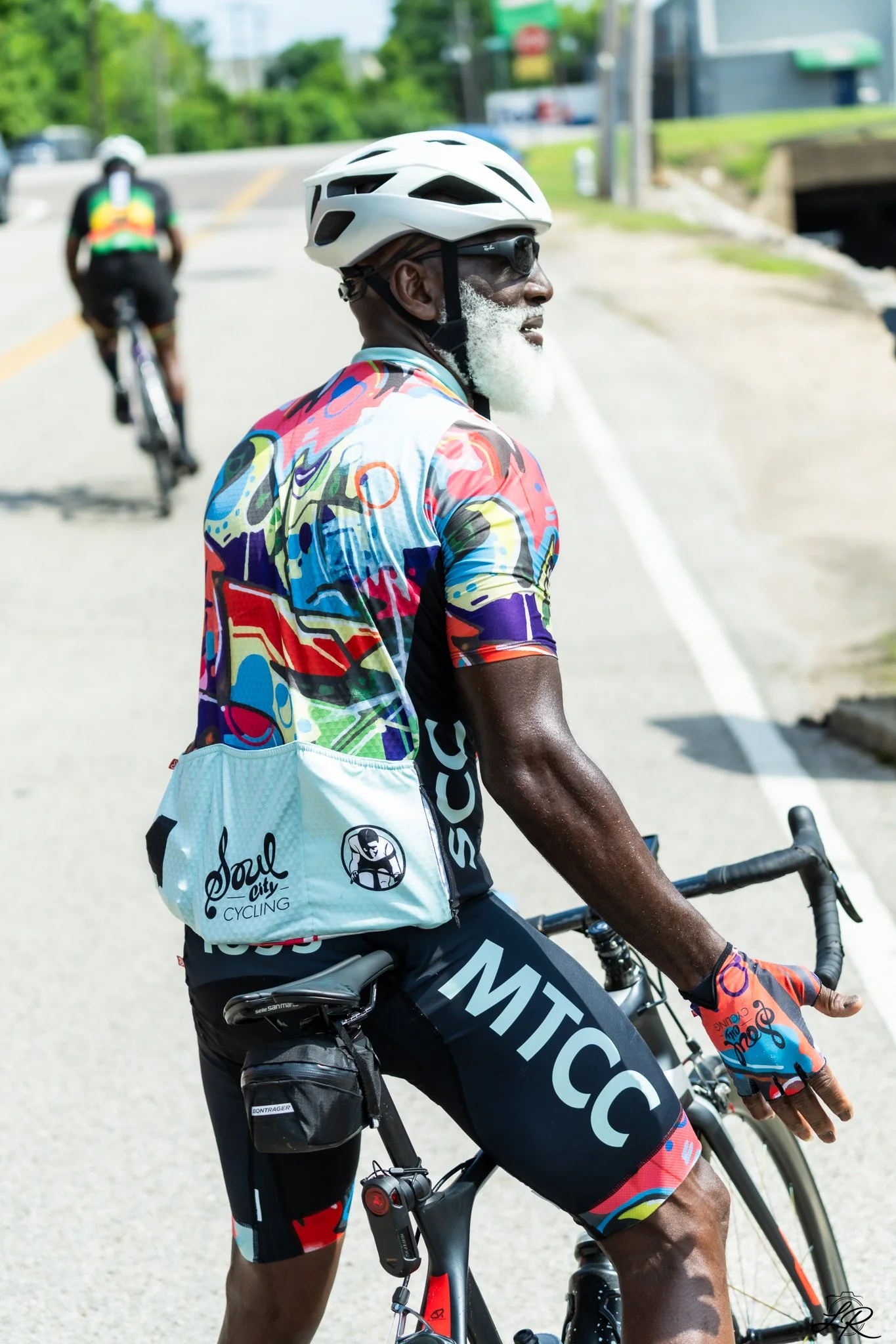 Man riding bike wearing a white helmet, colorful cycling shirt, and sunglasses, with a cyclist in the background on a sunny day.
