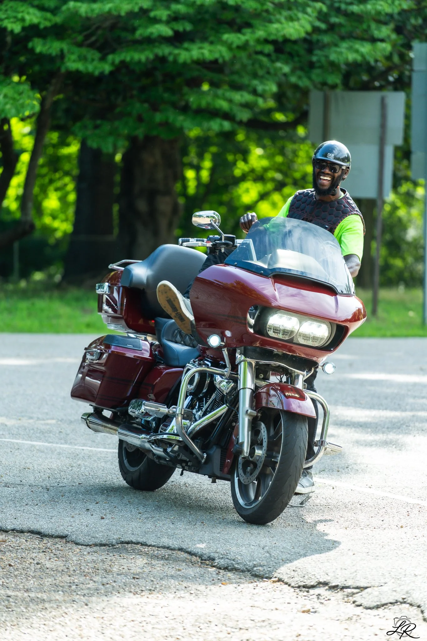 A man wearing a helmet, sunglasses, and a black vest riding a red motorcycle on a paved road outdoors with green trees in the background, smiling.