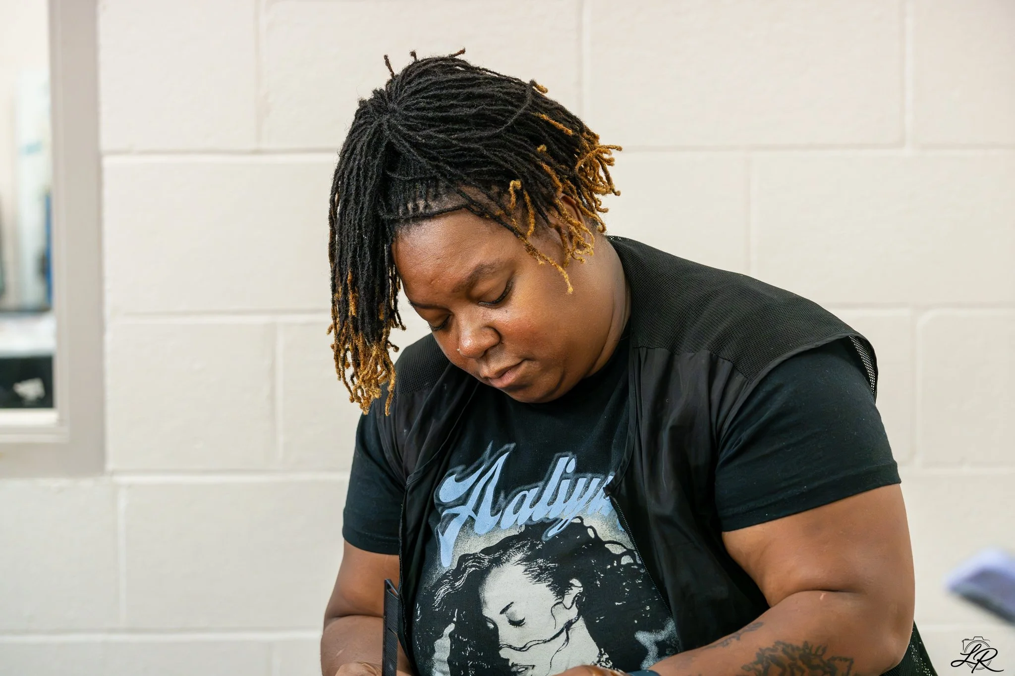 A woman with dreadlocks, some light brown, wearing a black T-shirt with a black leather vest, looking down, inside a room with white cinderblock walls.