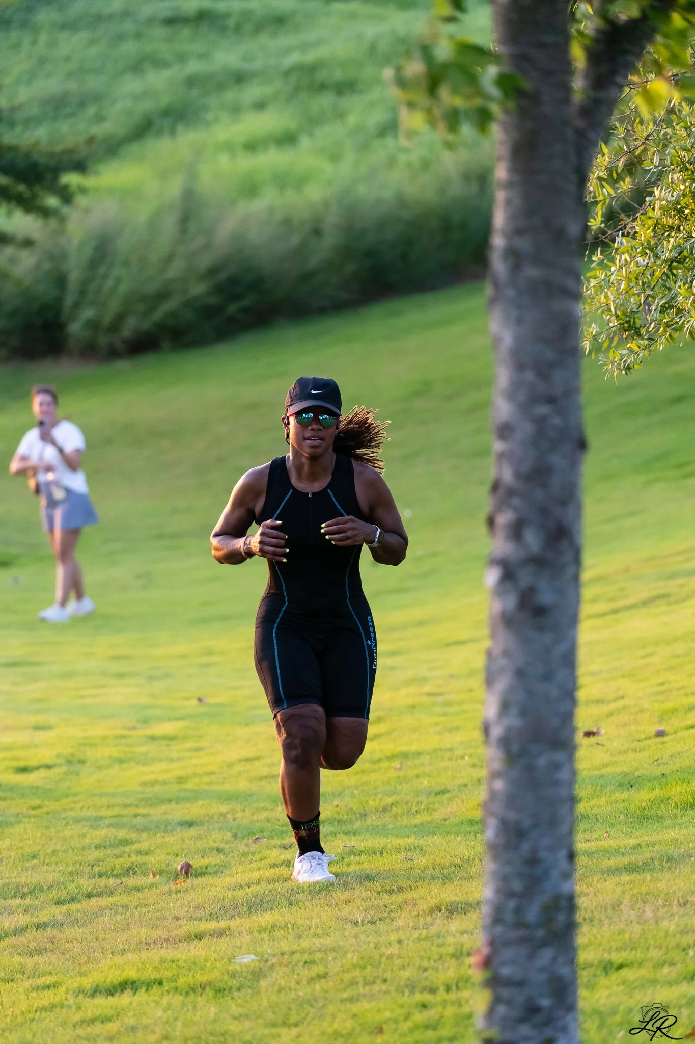 A woman running outdoors on a grassy hill, wearing a black athletic outfit, sunglasses, a black cap, and a fitness tracker, with trees and a person in the background.
