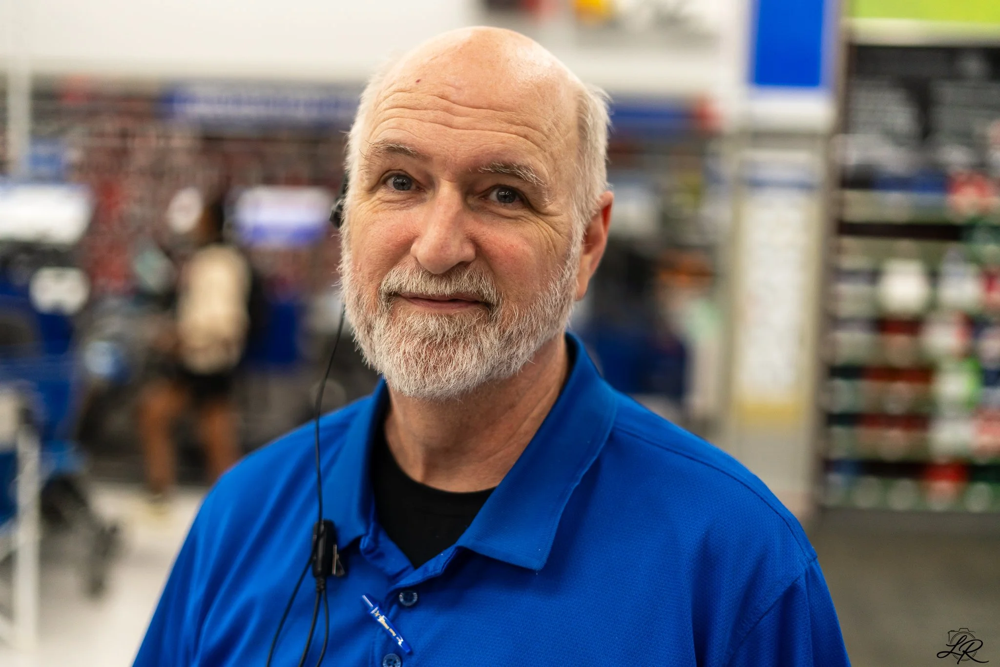 Close-up of a smiling older man with a beard and bald head, wearing a blue shirt, in a store aisle with shelves of products in the background.