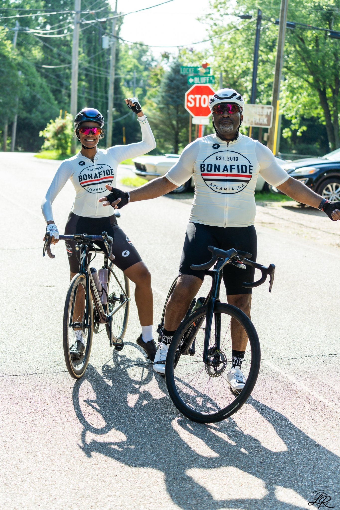 Two cyclists wearing white jerseys with 'Bonafide Atlanta, GA' printed on them, wearing helmets and sunglasses, standing with their bikes on a sunny street, casting shadows on the pavement with trees and stop signs in the background.