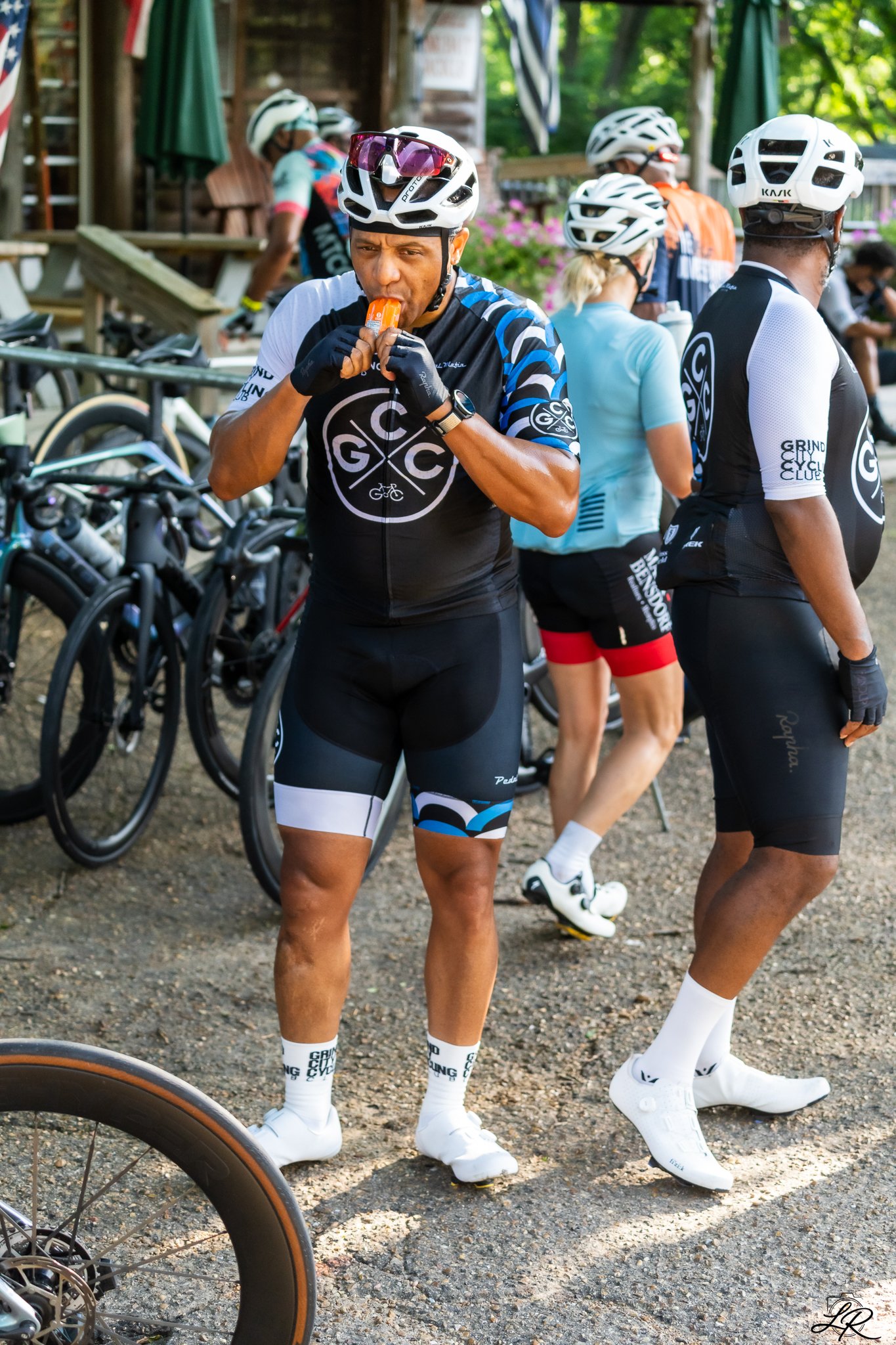 A group of cyclists taking a break outdoors, some are wearing black and white cycling jerseys and helmets, with one man in the foreground eating an orange popsicle, others are standing around with bikes in the background, and a partial view of a bicy