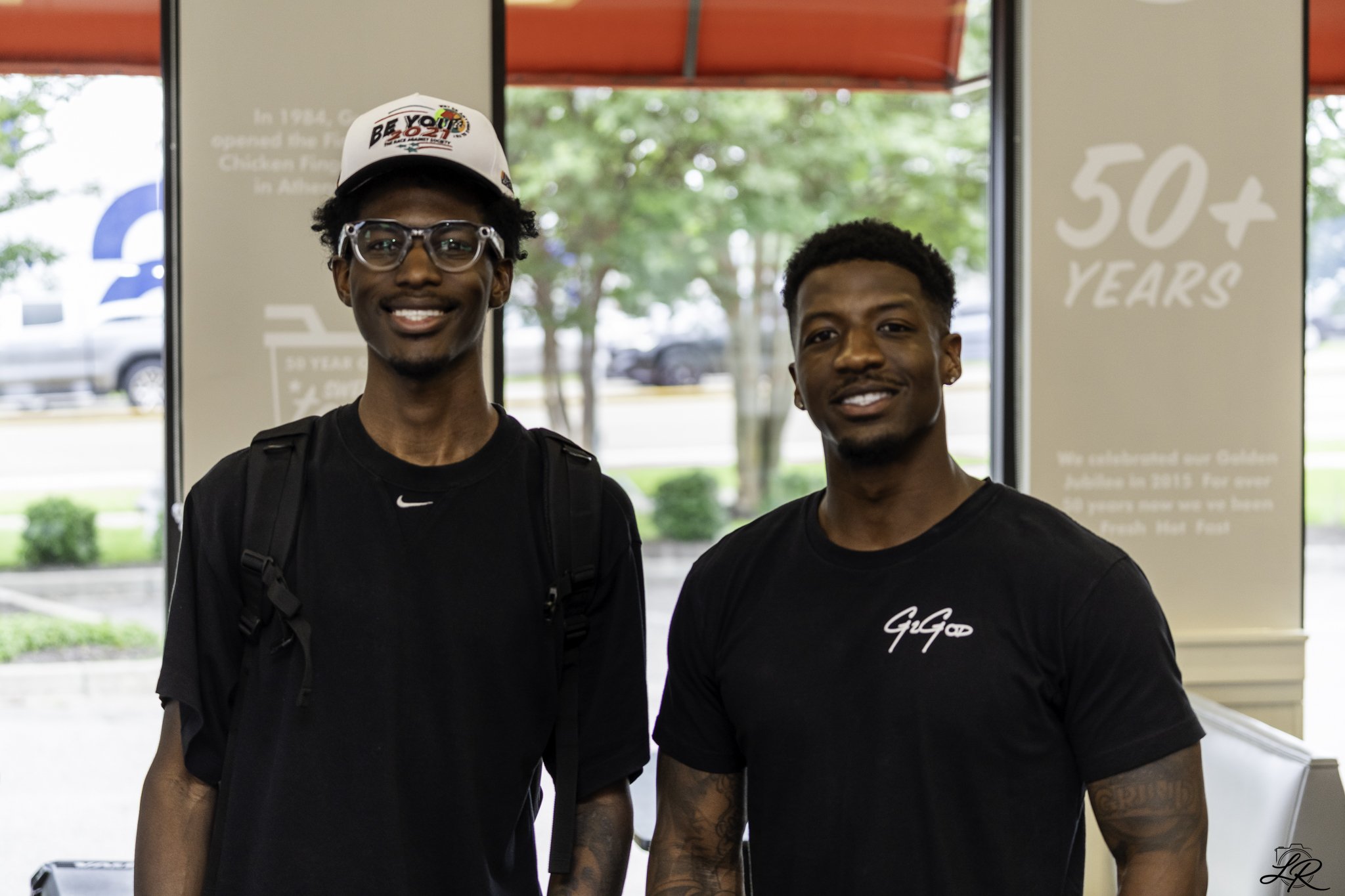 Two young men standing indoors, smiling at the camera. The man on the left wears a white cap with colorful text and logos, glasses, and a black hoodie with a backpack. The man on the right has short curly hair, tattoos on his arm, and wears a black T