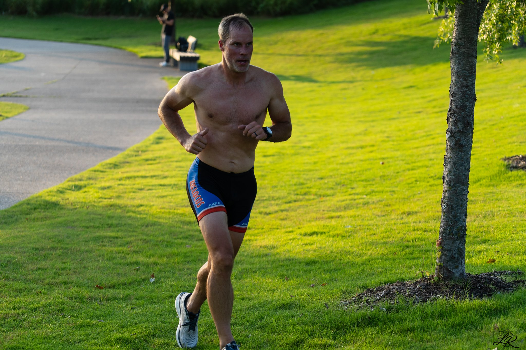 A shirtless man running on a grassy path in a park, with a woman in the background using a phone. The man is wearing a black sports watch, black and blue shorts, and running shoes, with sunlight casting a glow on the scene.