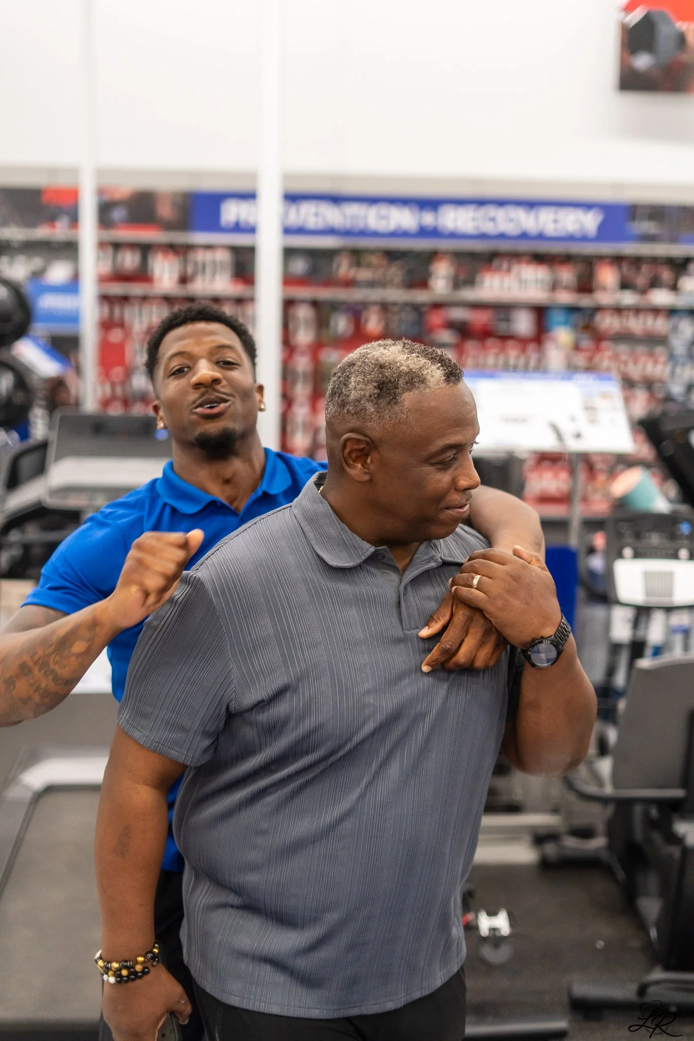 Two men embracing inside a store, with shelves and signs in the background. One man is in a gray shirt and the other in a blue shirt.