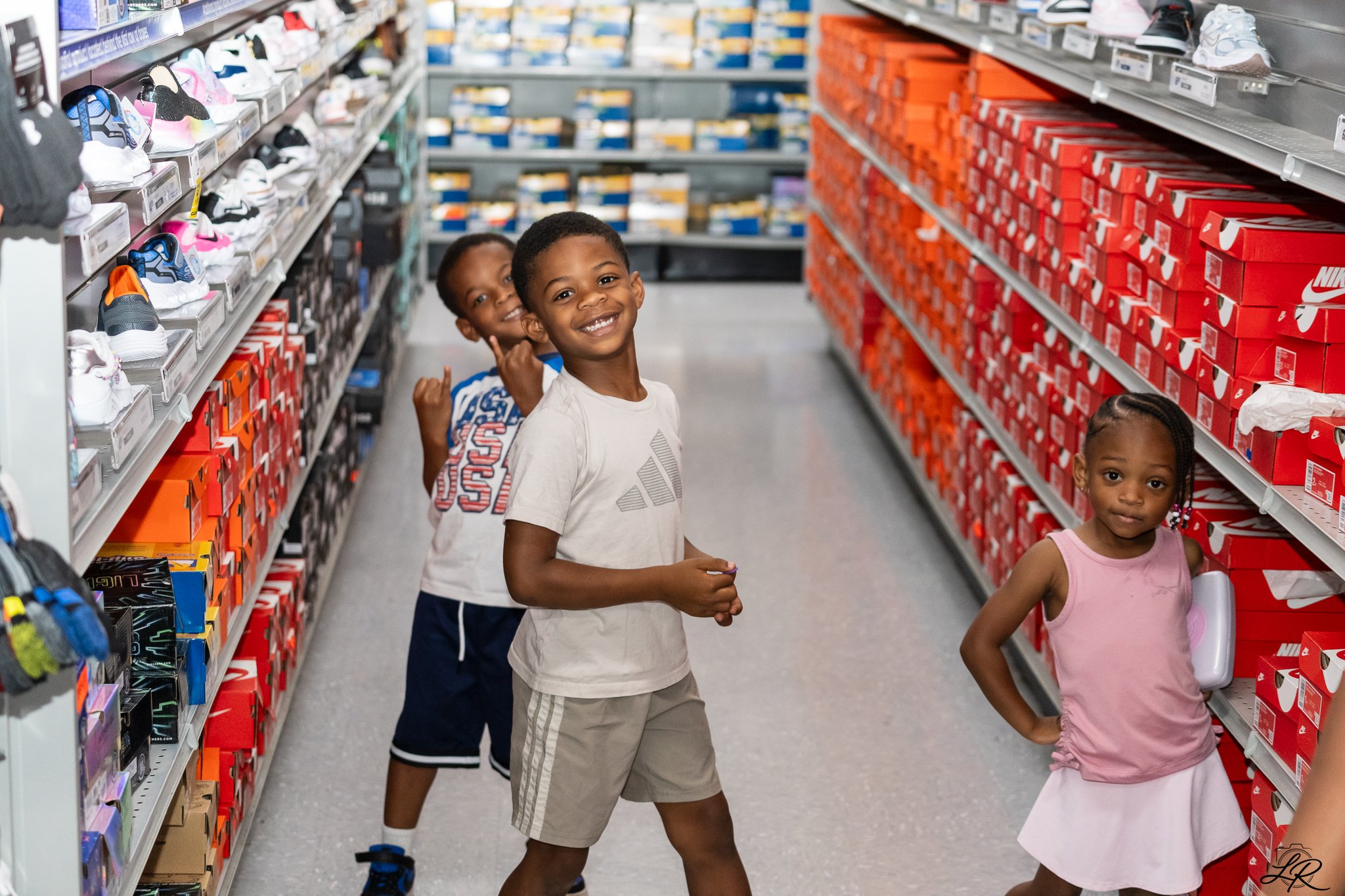Three children shopping for shoes in a store aisle with shelves full of sneaker boxes.