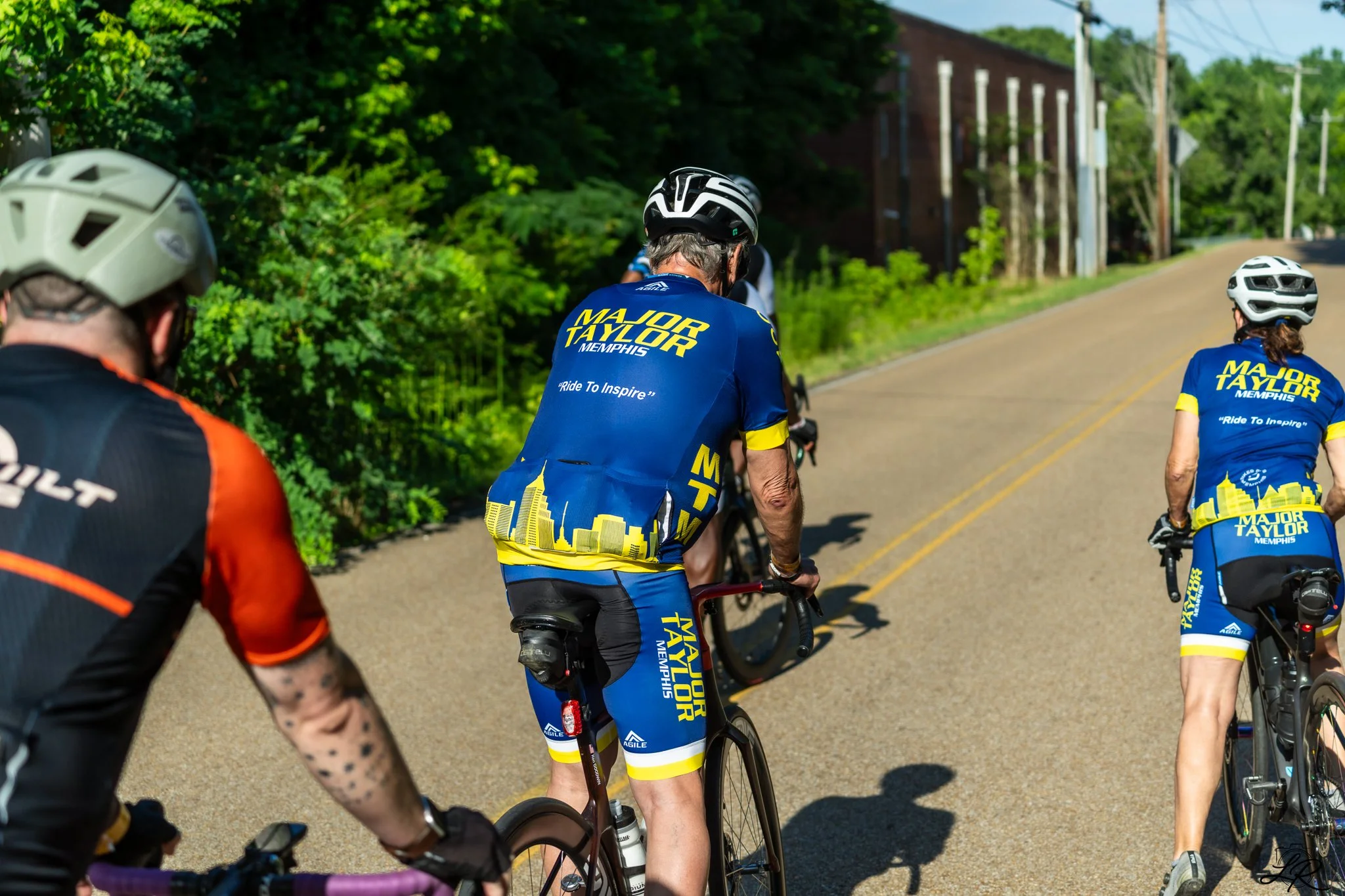 Cyclists riding on a rural road with green trees and buildings in the background, wearing colorful jerseys and helmets.