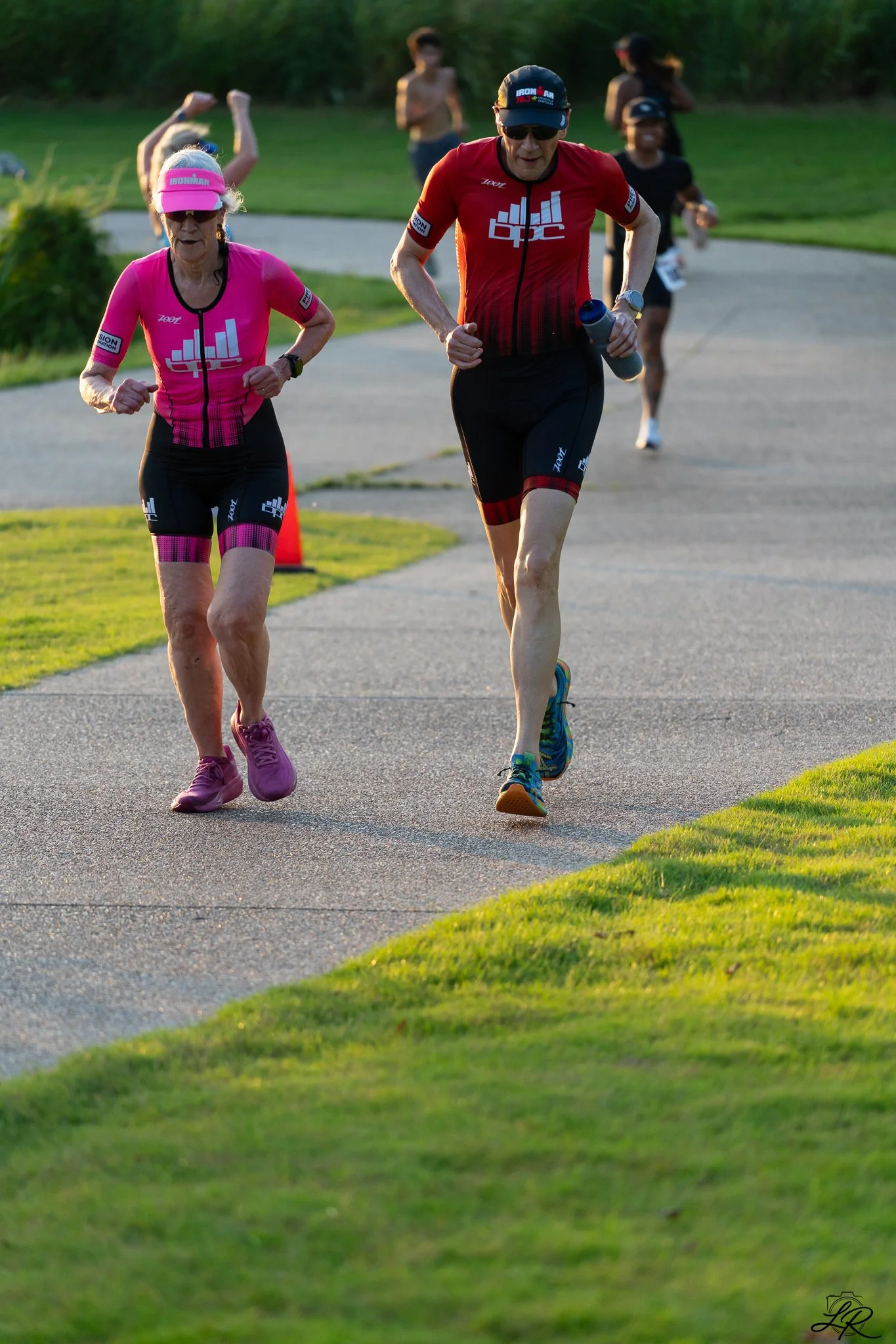 A group of runners participating in a race outdoor, with two leading female runners wearing pink and black and a male runner in red shirt and black shorts, on a paved trail with green grass and trees in the background.