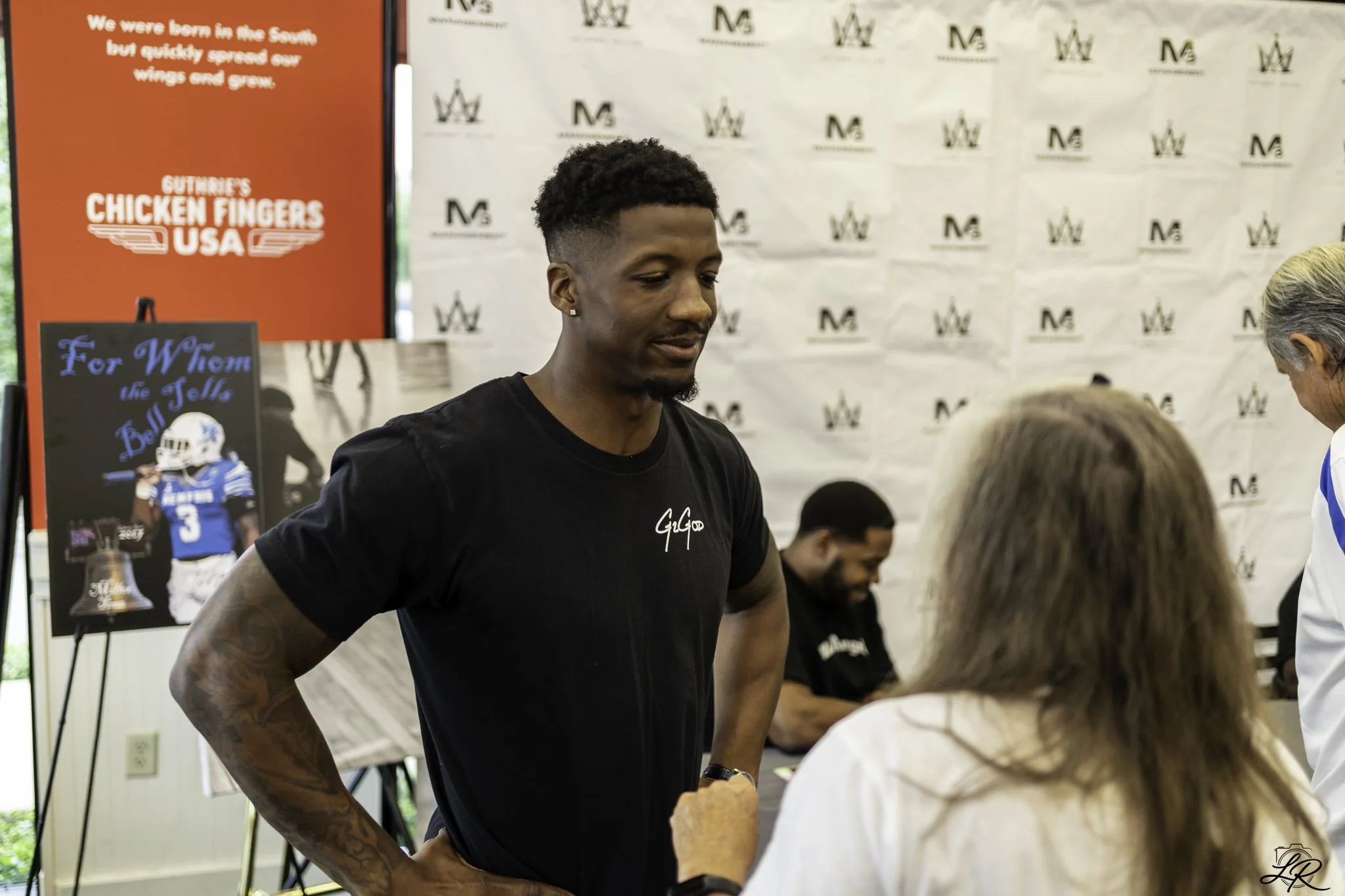 A young man with short curly hair and tattoos on his arms is standing indoors, wearing a black T-shirt, and engaging in conversation with multiple people. The background features a white banner with black logos and a red display with white text about