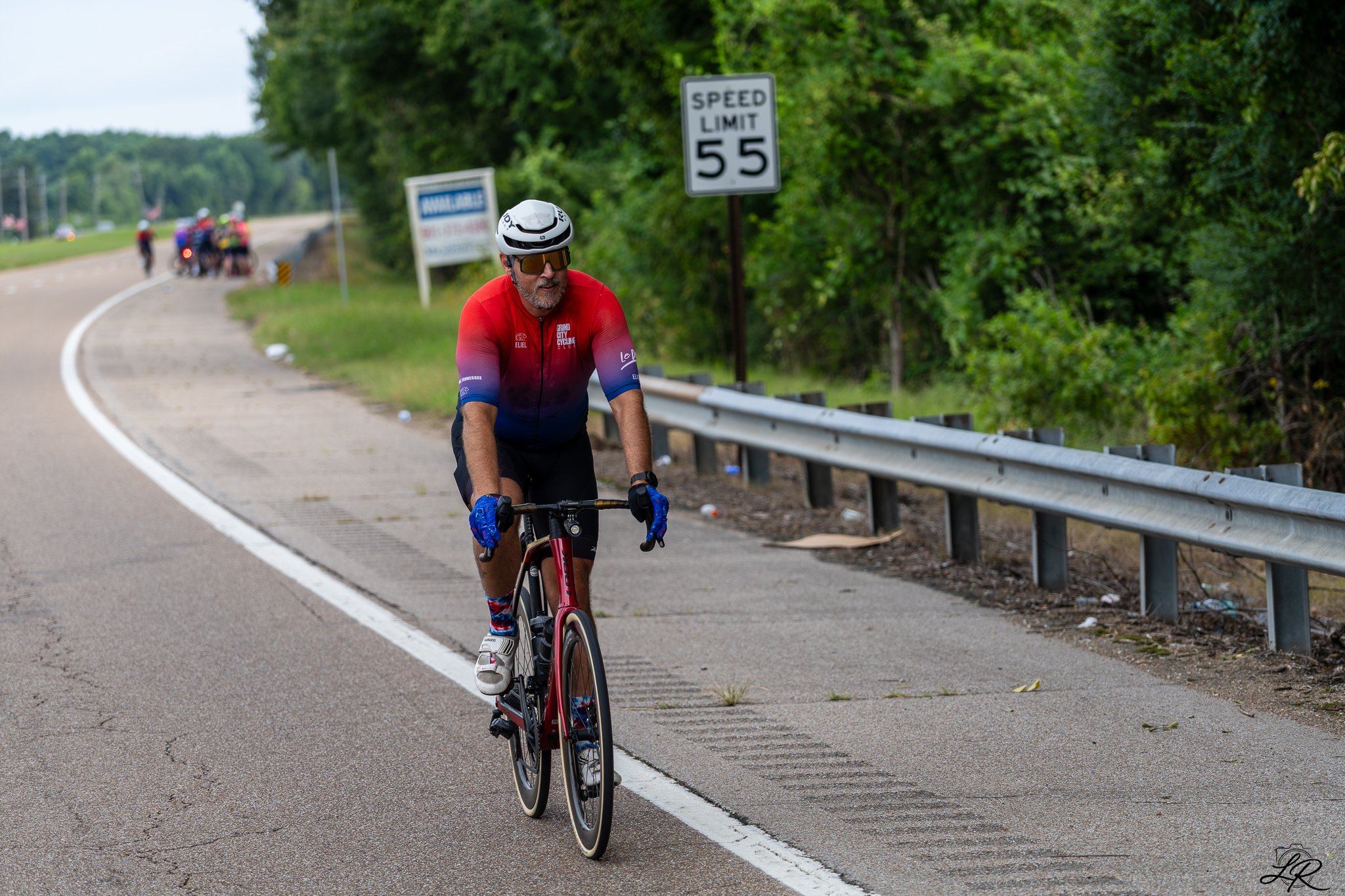 A man in a red cycling jersey and a white helmet riding a red bicycle on the side of a road, with a speed limit sign showing 55 mph and a group of cyclists in the background.