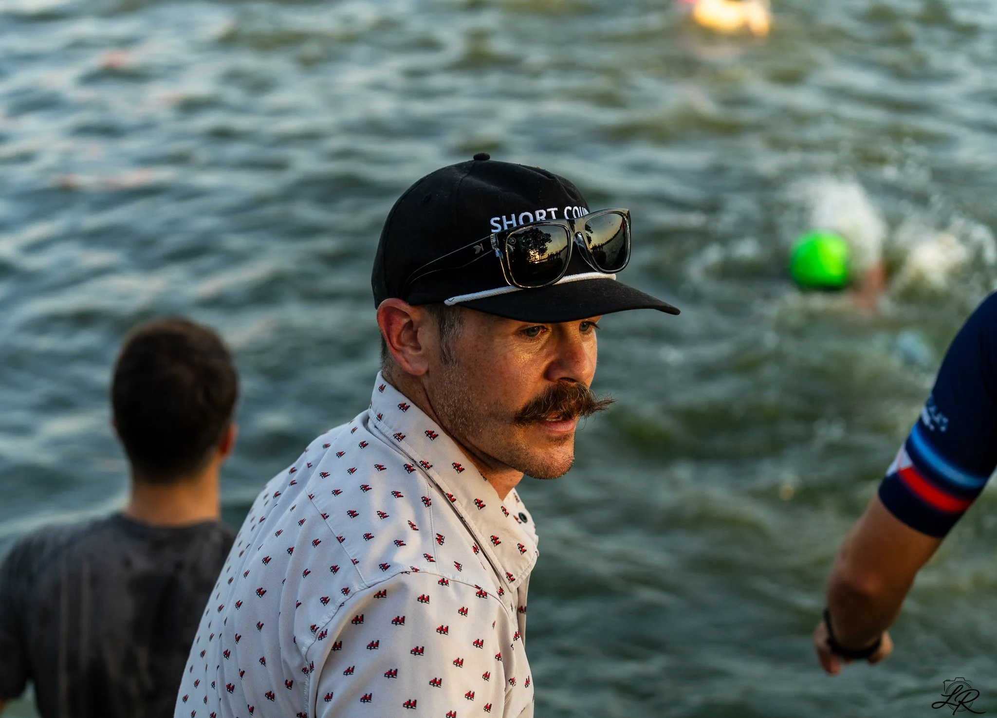 A man with a mustache and goatee wearing a white shirt with small red and black patterns, a black cap with the word 'SHORT COURSE', and sunglasses perched on the cap, standing near water. The background shows rippling water and other indistinct figur