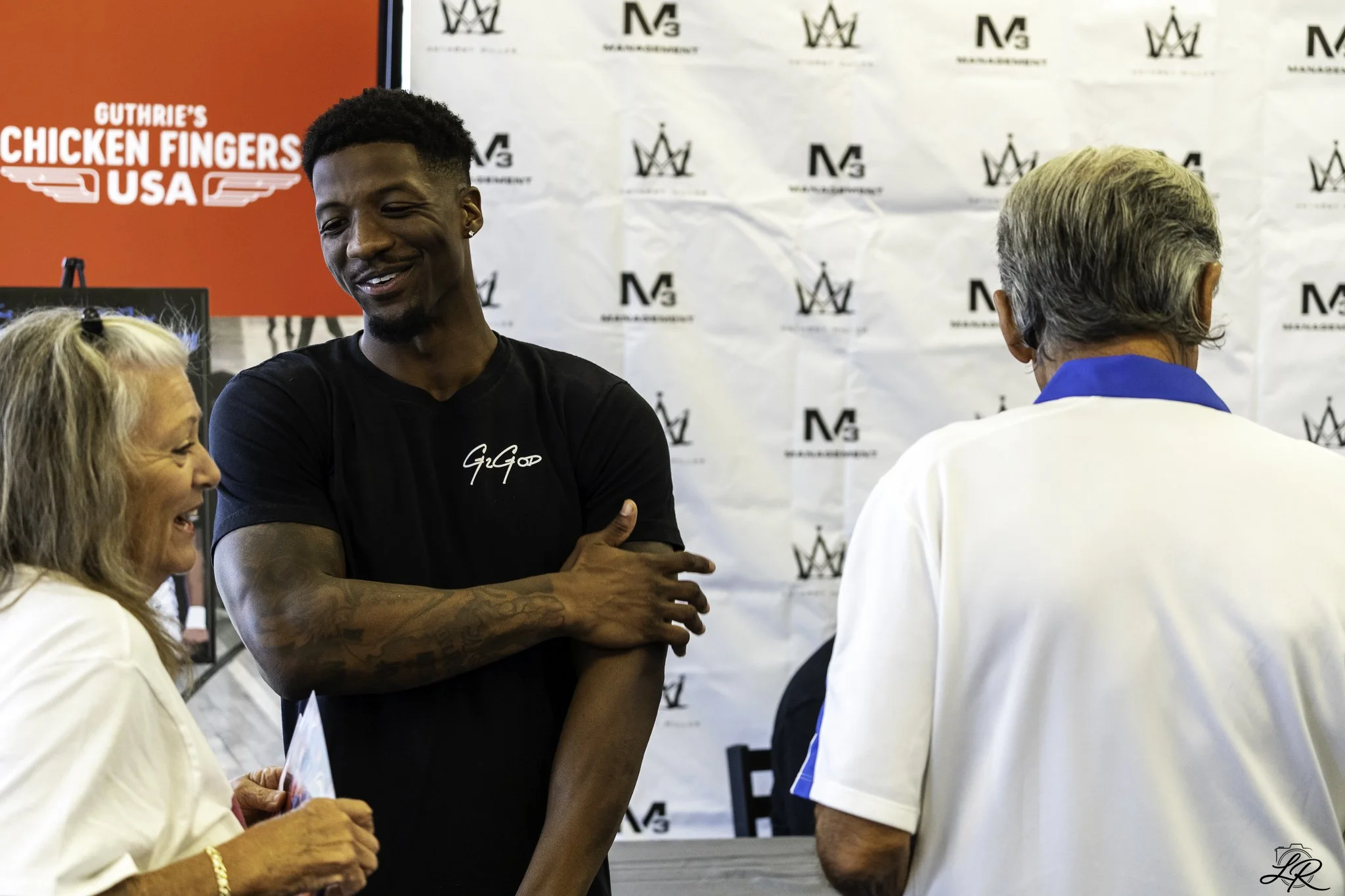 A young man smiling and talking with two women at an event, with a red and white background displaying the logos and a sign that reads 'Guthrie's Chicken Fingers USA'.