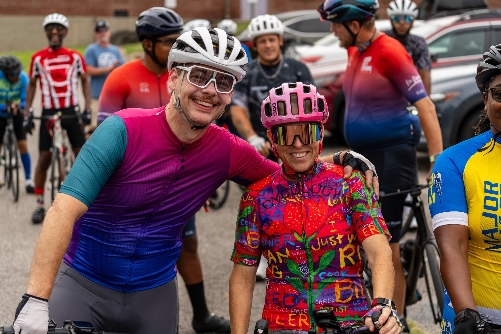 A group of cyclists dressed in colorful cycling gear, including helmets and sunglasses, gathered outdoors. Two cyclists are smiling at the camera, with one wearing a pink helmet and a rainbow-colored jersey, and the other in a white helmet and pink a