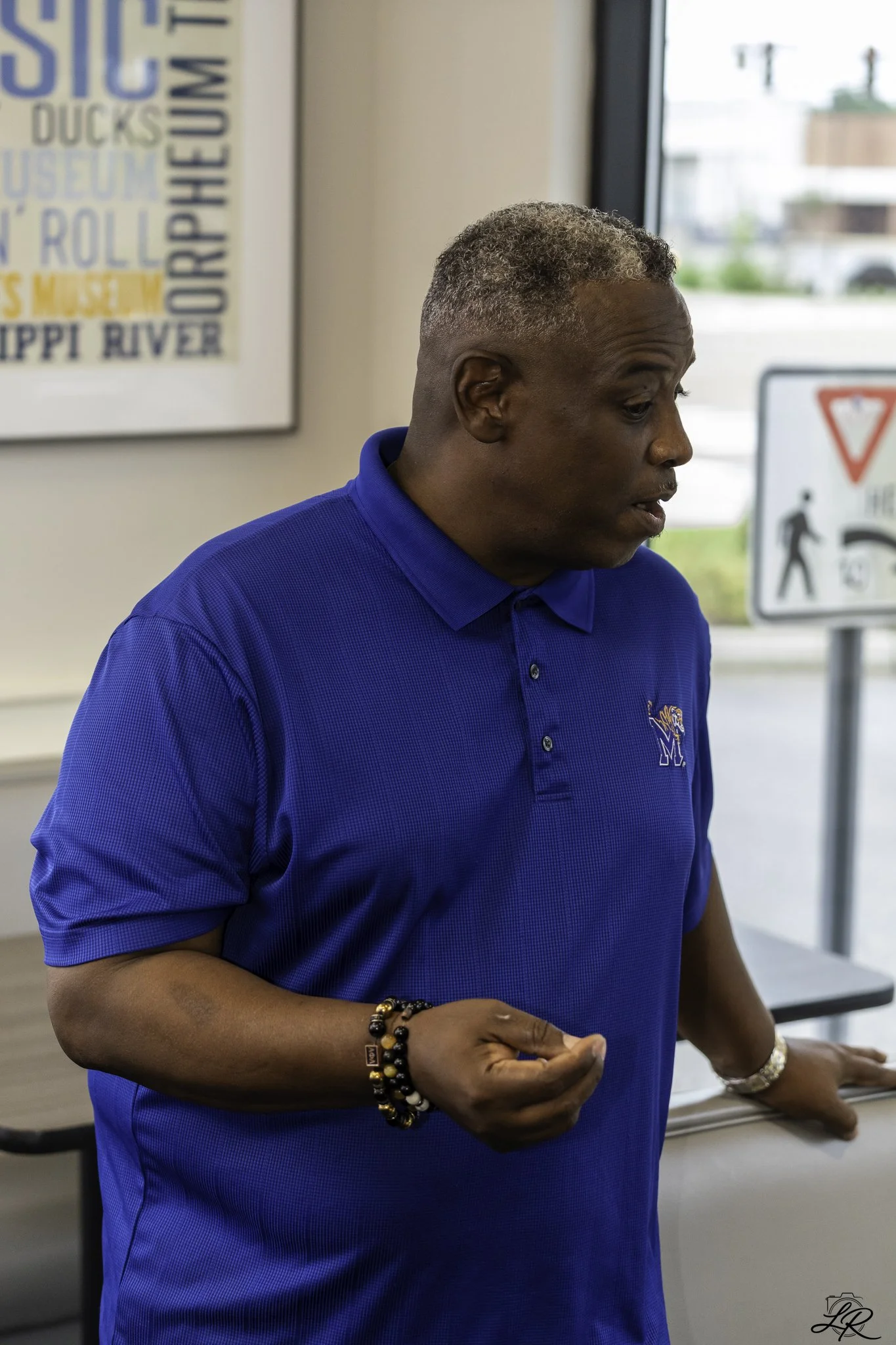 A middle-aged man with short, curly gray hair wearing a blue polo shirt, standing indoors near a window with signs and posters in the background.