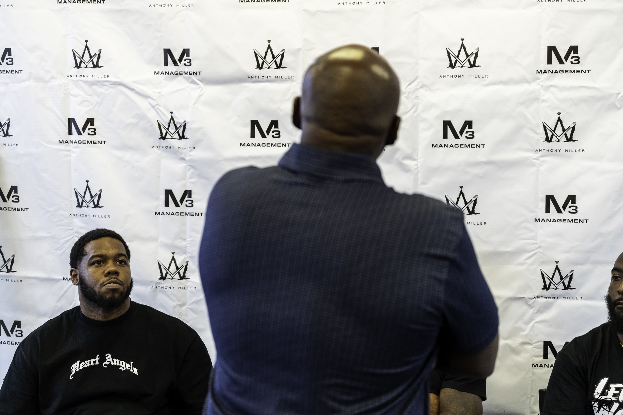 Three men seated in front of a white backdrop with logos for Anthony Miller and Management. The man in the center is facing away, with a shaved head, wearing a dark suit. The man on the left has short hair, a beard, and is wearing a black T-shirt tha