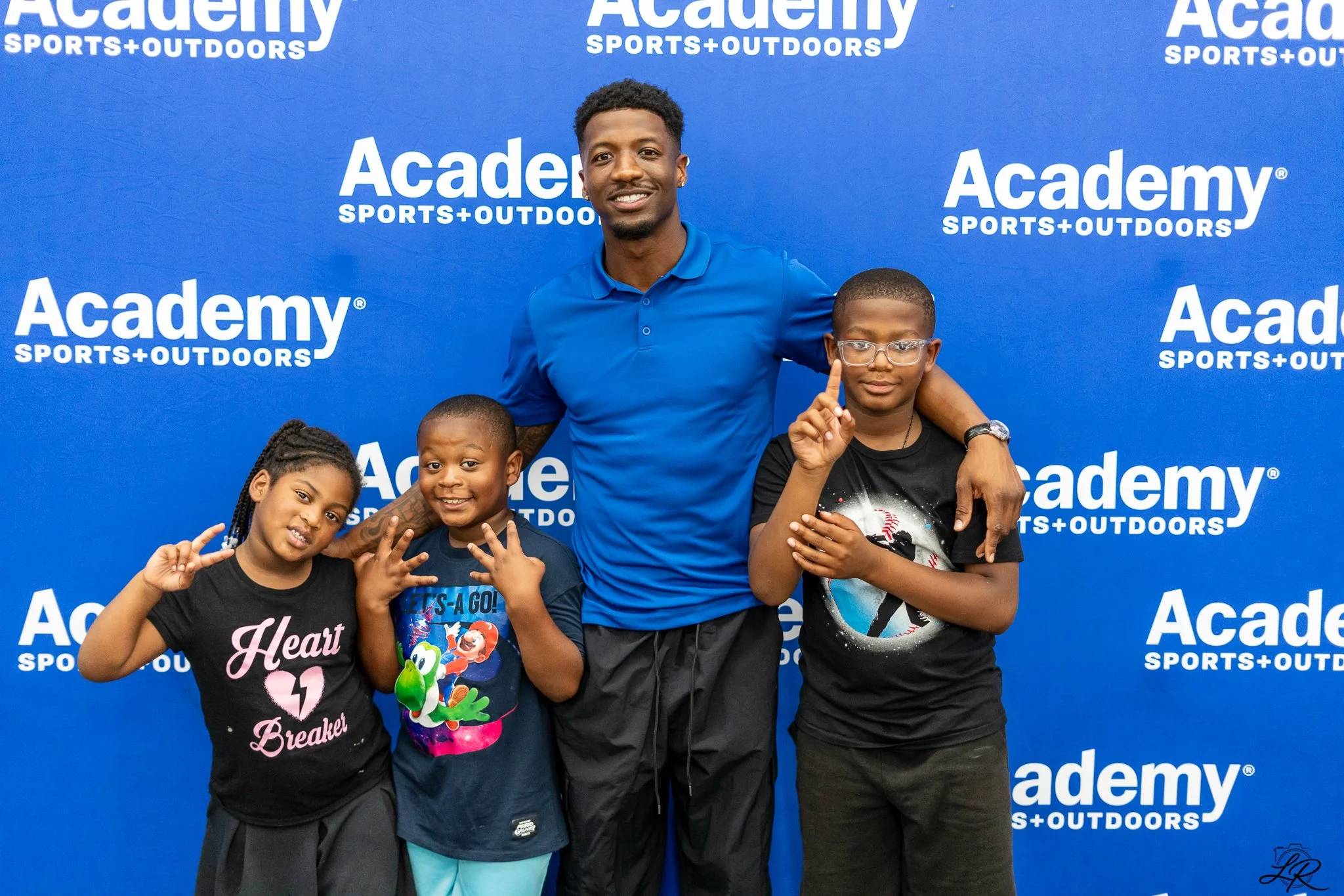 A man with three children posing in front of a blue backdrop with white 'Academy Sports + Outdoors' logos. The man is smiling, wearing a blue polo shirt, and has his arm around one child. The children are making hand gestures, and one is wearing glas