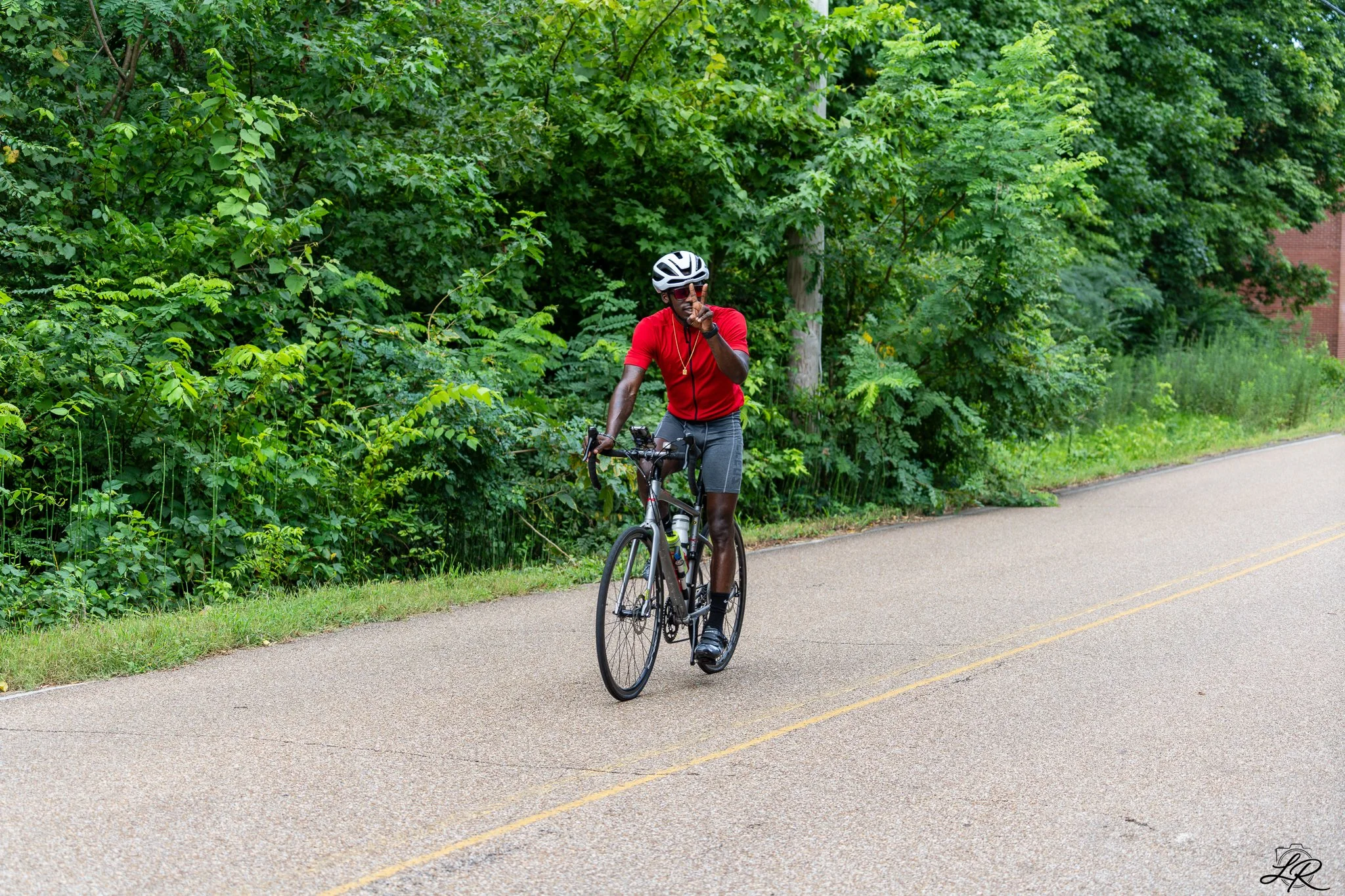 A man wearing a red shirt, gray shorts, and a bicycle helmet rides a bicycle on a paved road with double yellow lines, next to a dense green bush and trees.