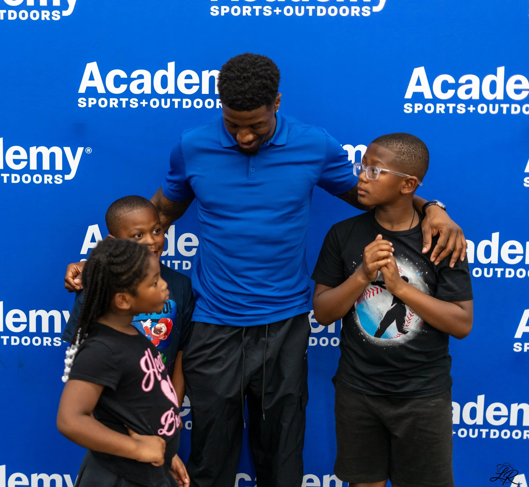 A woman with braided hair and a black t-shirt with pink text, a man in a blue polo shirt, and a young girl with glasses and a gray t-shirt with a baseball graphic are standing in front of a blue backdrop with white text that reads 'Academy SPORTS + OUTDOORS.'