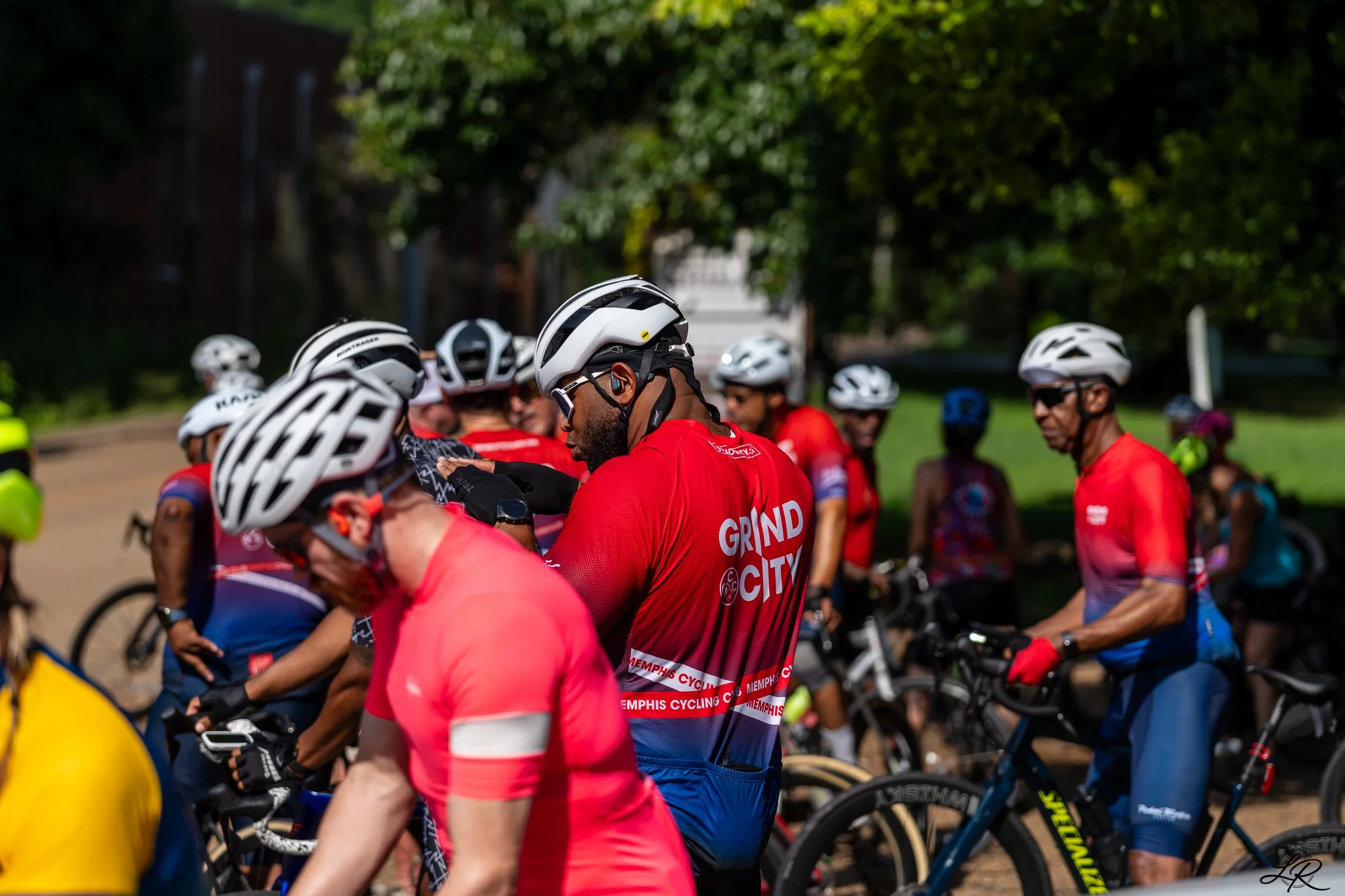 Group of cyclists wearing helmets and red shirts gather outdoors on a sunny day with green trees in the background.