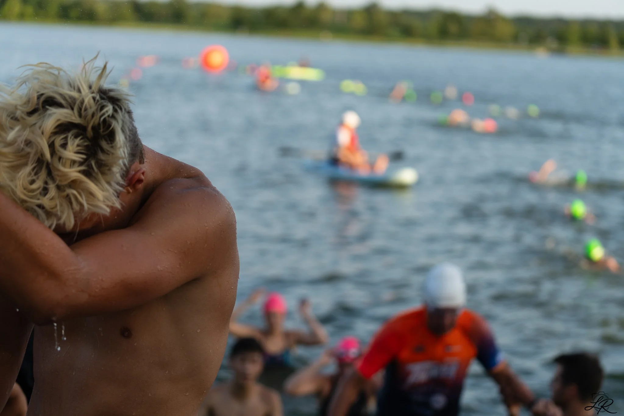 A person hugging a blonde boy on a dock near a lake with people swimming and floating on the water in the background.