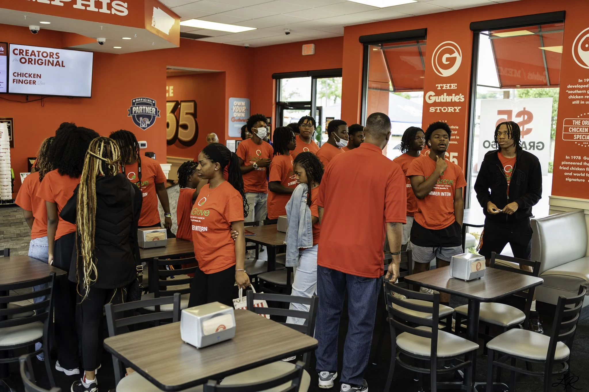 Interior of a fast-food restaurant with a group of mostly young people wearing red shirts, some wearing face masks, waiting in line or standing around. Walls are decorated with various posters and signs, including a large digital menu displaying food