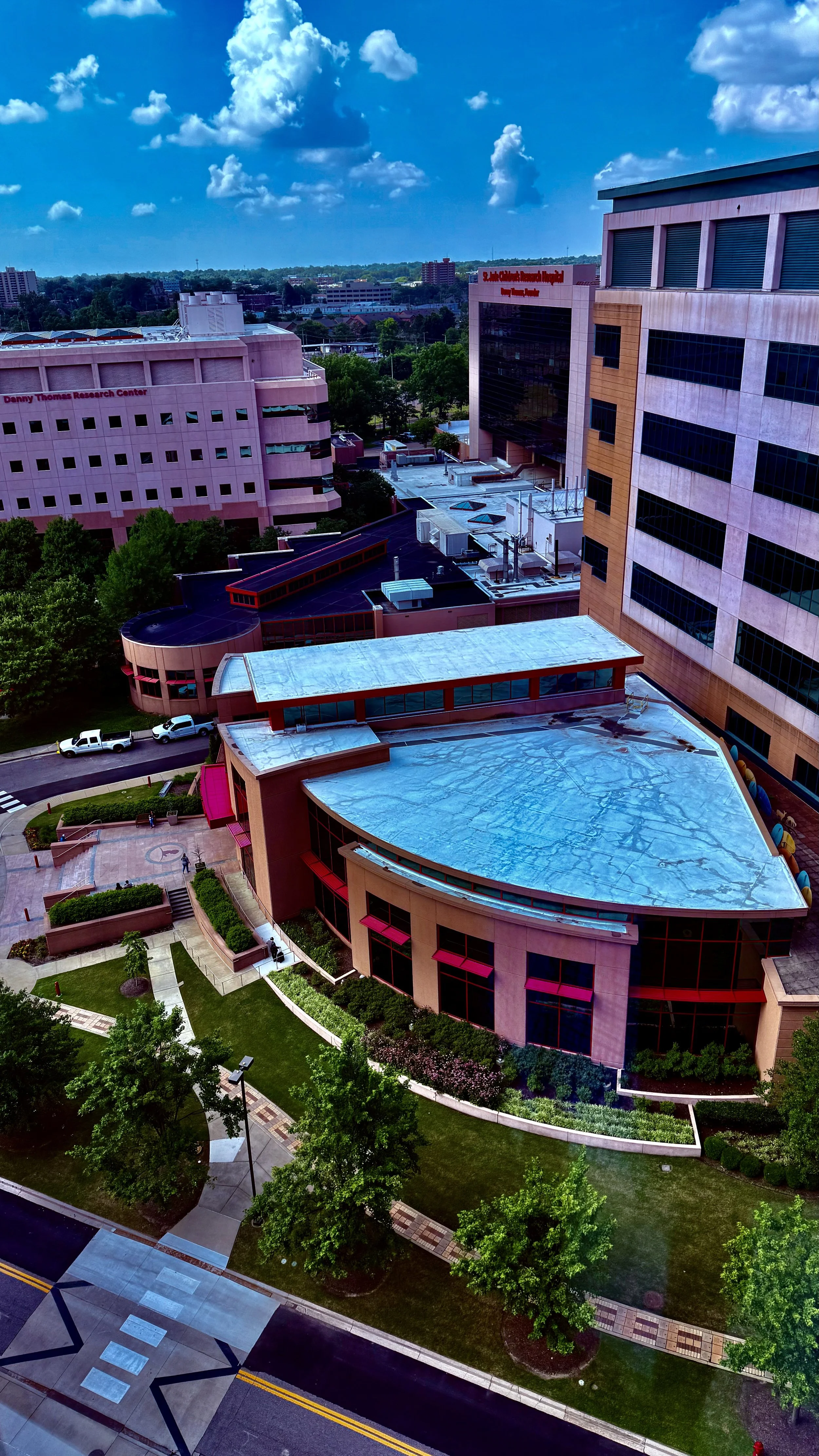 Aerial view of a cityscape showing office buildings, green trees, and a parking lot under a partly cloudy sky.