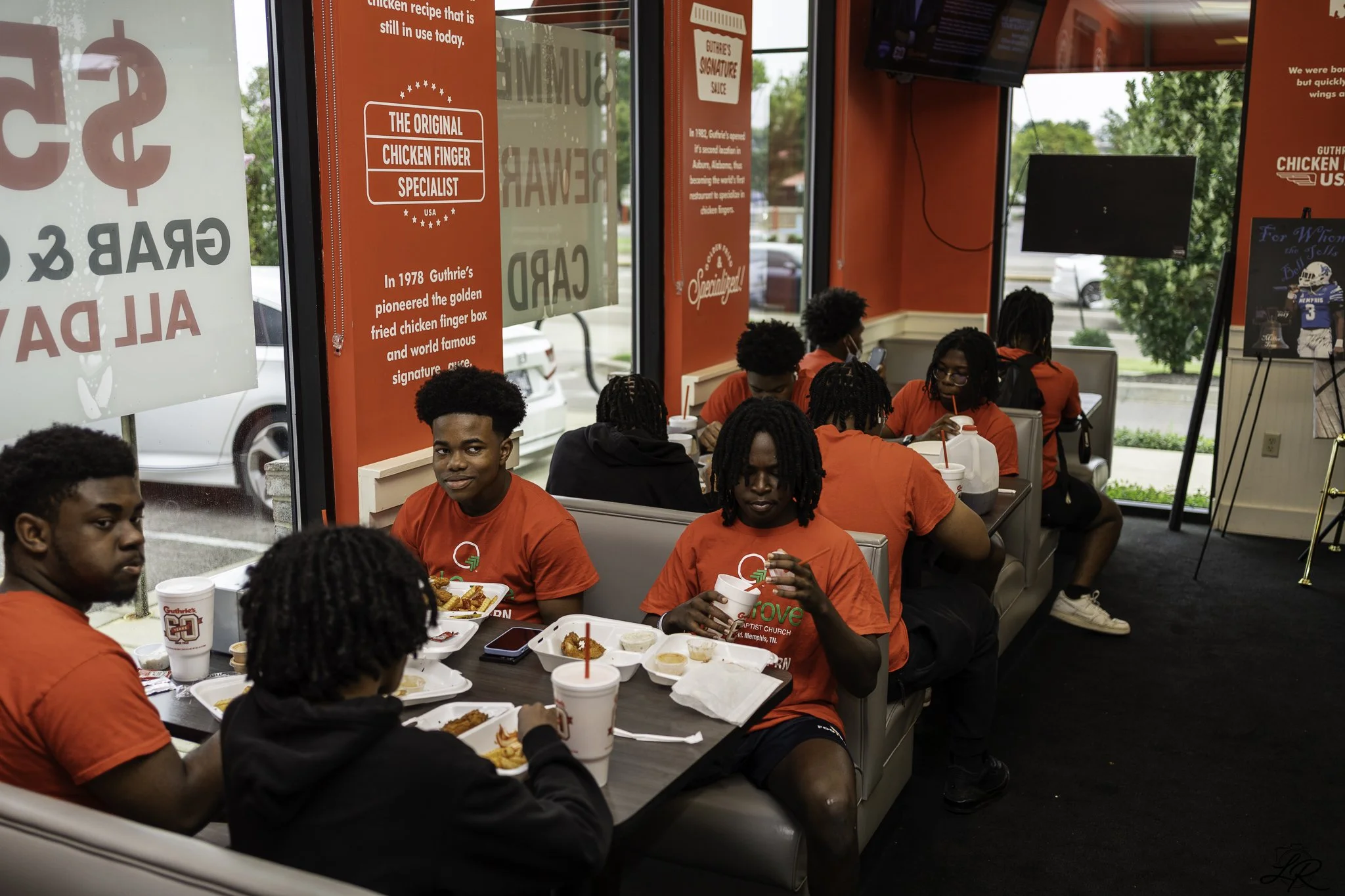 A group of young people sitting inside a Gutheries fast food restaurant, eating chicken and drinking beverages with red cups, many of them are wearing red shirts, with signs and posters about chicken on the windows and walls.