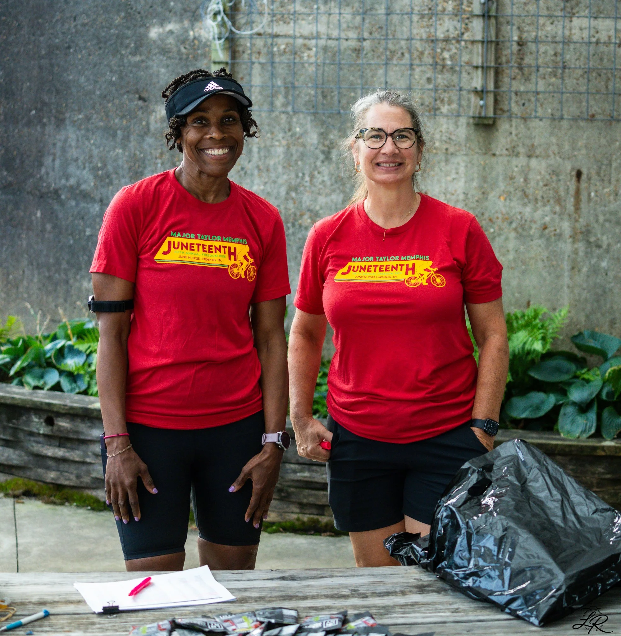 Two women wearing red t-shirts with a July 2025 Juneteenth motorcycle event logo, standing outdoors behind a table with papers and writing instruments, smiling at the camera.