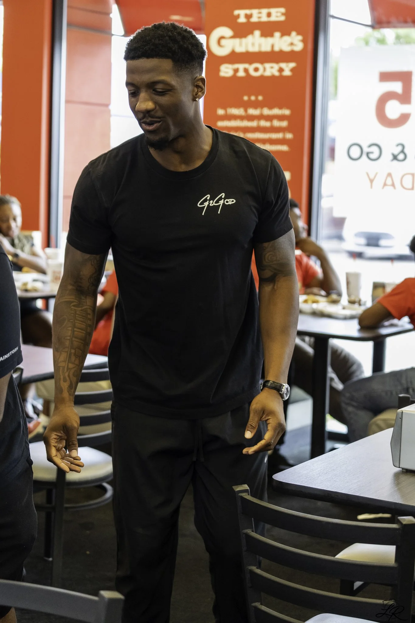 A man wearing a black t-shirt with white writing on it, standing inside a fast food restaurant with bright red walls and signage, surrounded by other patrons seated at tables.