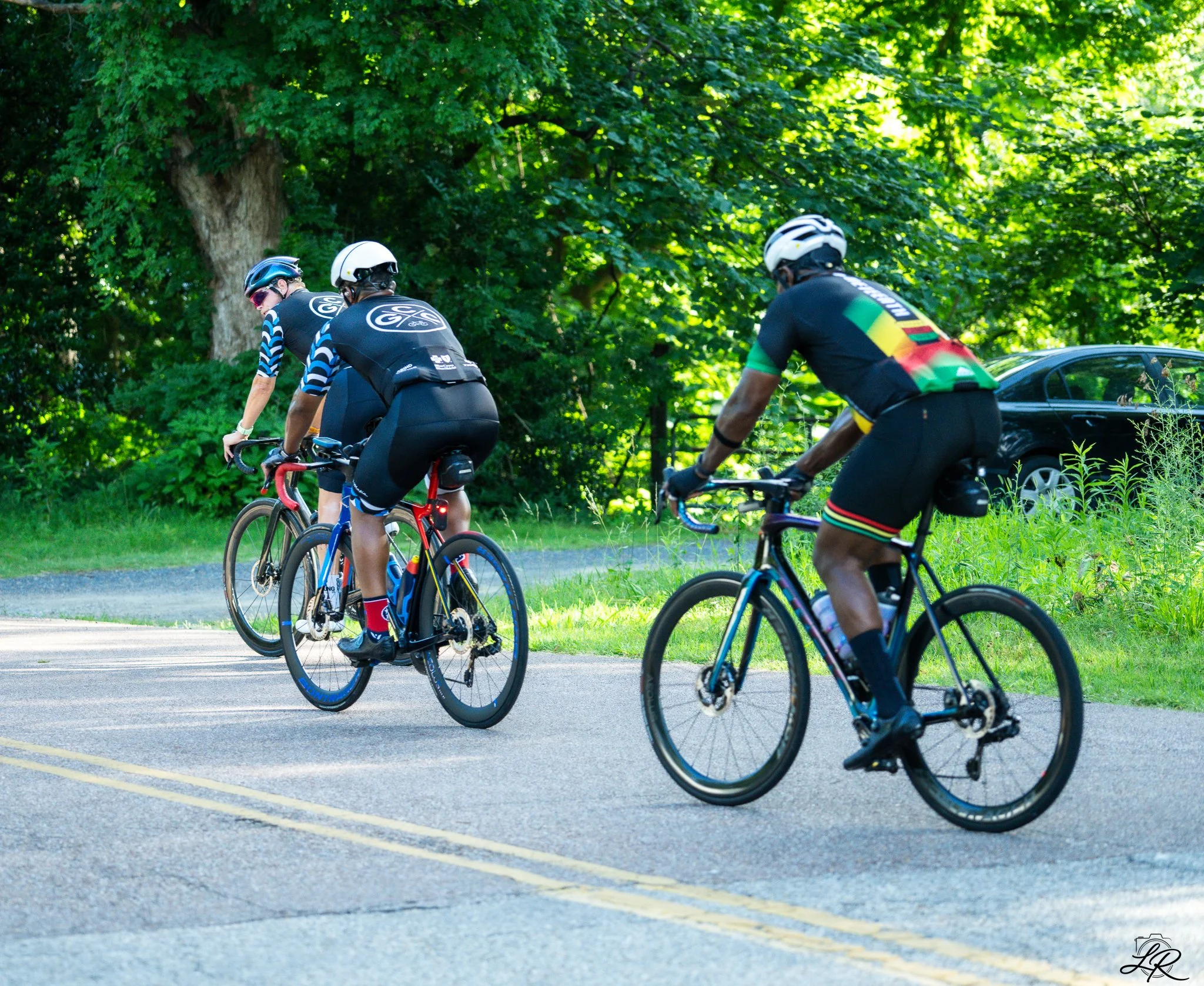 Three cyclists ride on a road surrounded by green trees, wearing helmets and sporty clothing.