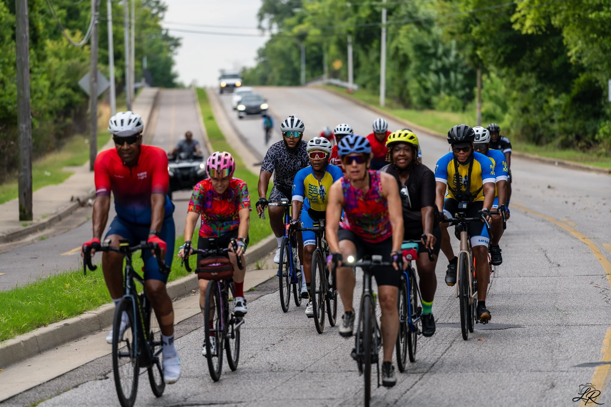 Group of cyclists riding on a road surrounded by green trees, with cars in the background.