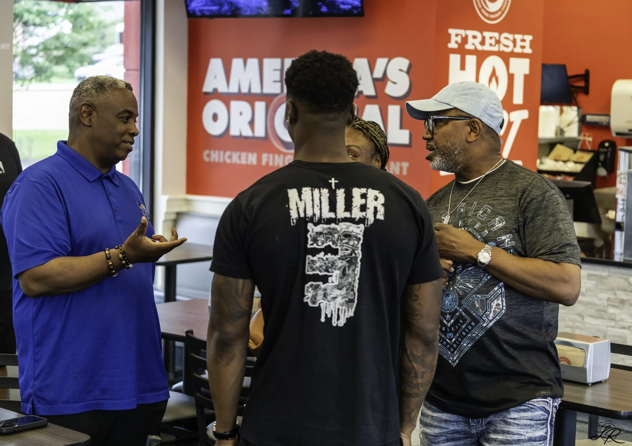 Four people engaged in conversation inside a fast food restaurant with a red and white branding background. One person is wearing a bright blue polo shirt, another has a black T-shirt with white graphics, the third in a gray cap and glasses, and a wo