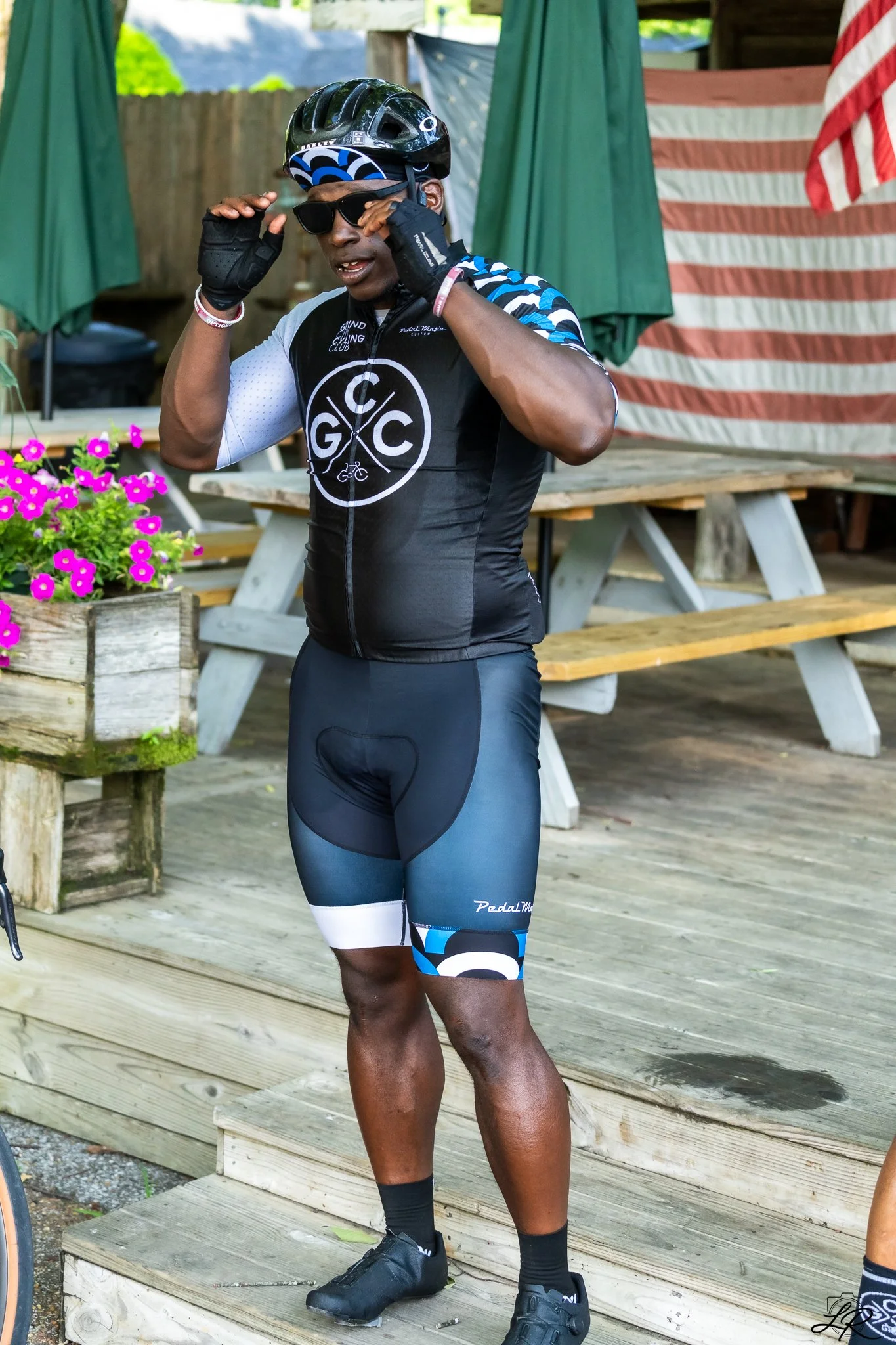 A male cyclist adjusting sunglasses, wearing a black and blue cycling jersey, shorts, helmet, and gloves, standing on a wooden deck with pink flowers in a planter nearby and an American flag in the background.