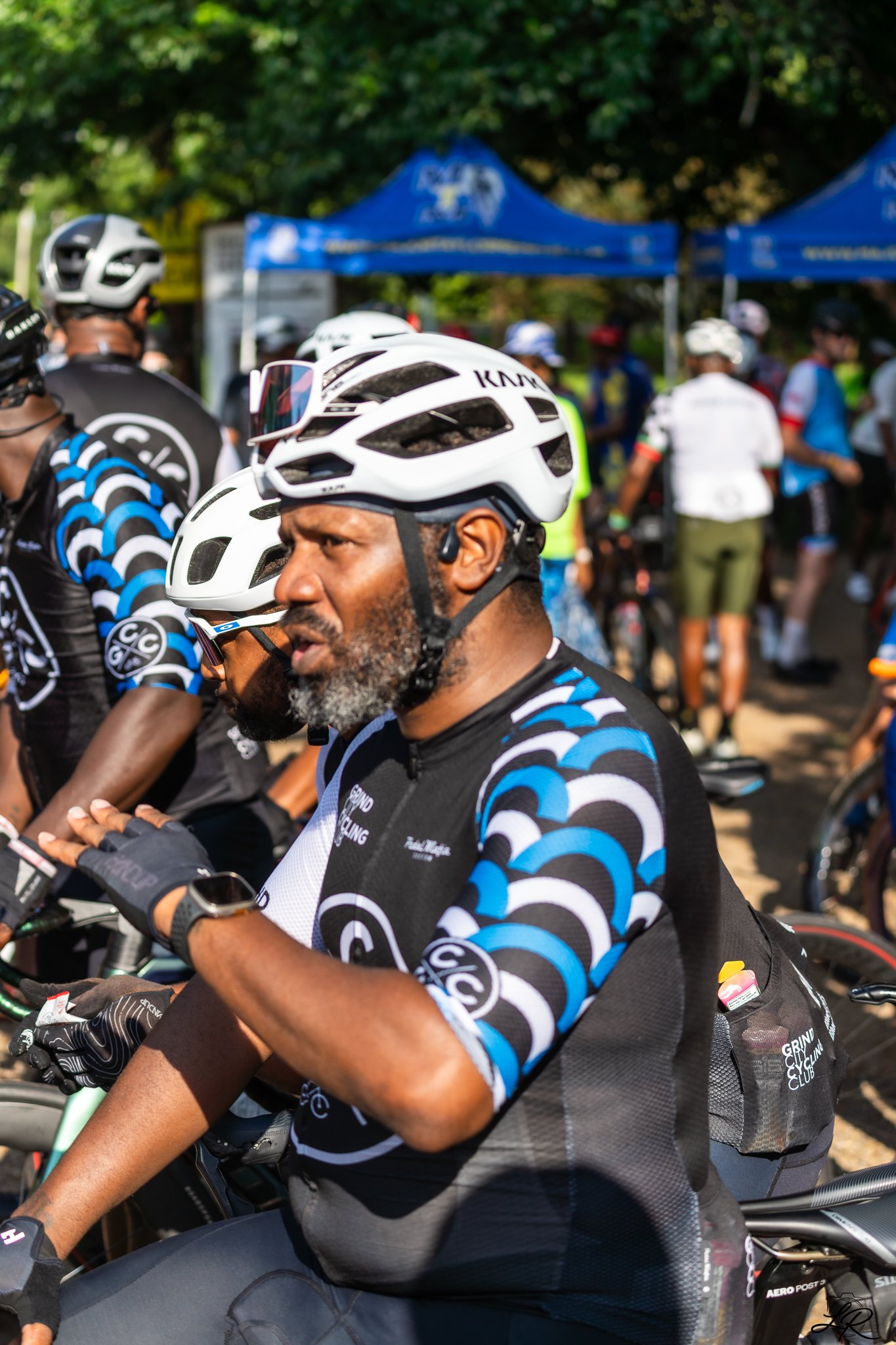 A group of cyclists wearing helmets and cycling jerseys gathered outdoors, with trees in the background and a blue canopy tent.