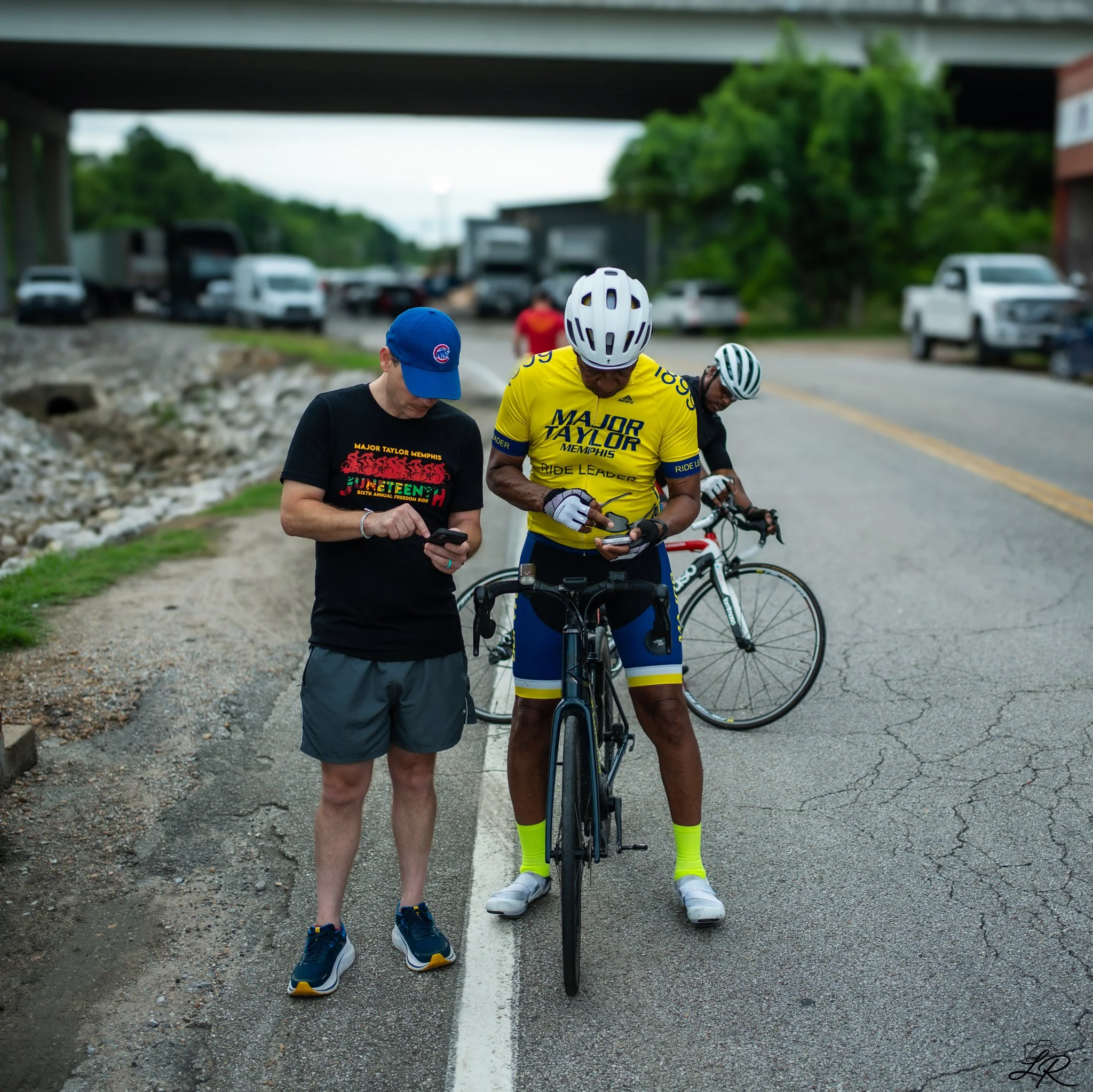 Three cyclists standing with their bikes on a rural road, looking at their phones, under a bridge, with parked cars and green trees in the background.