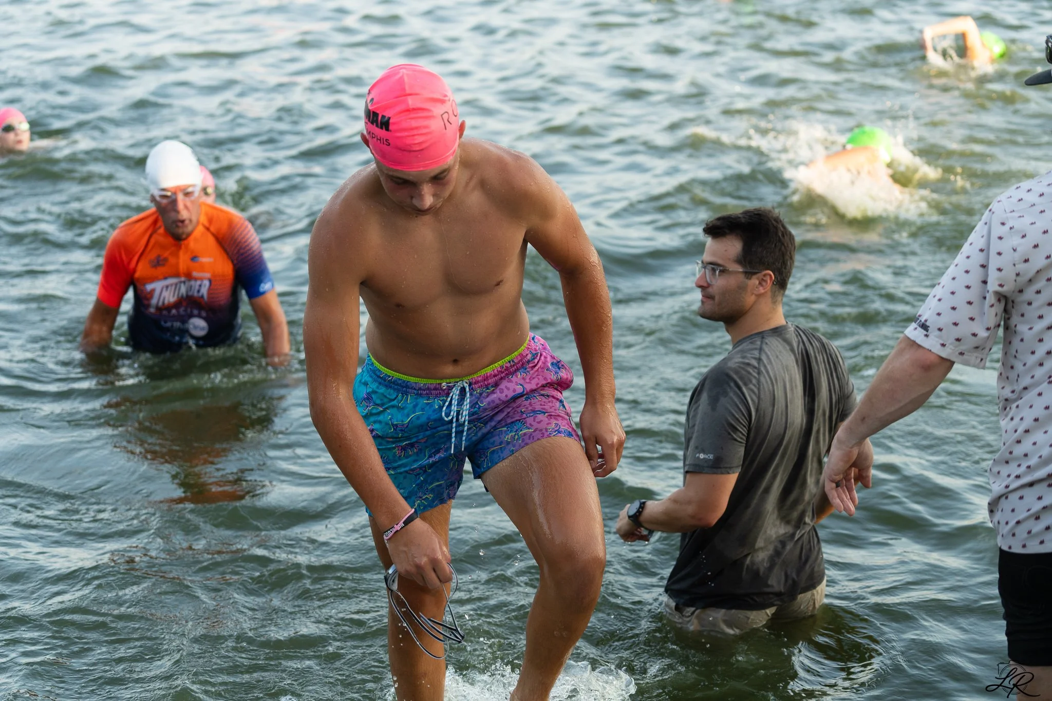 Male swimmer in colorful shorts walking out of the water during a triathlon, with other swimmers and event staff in the background.