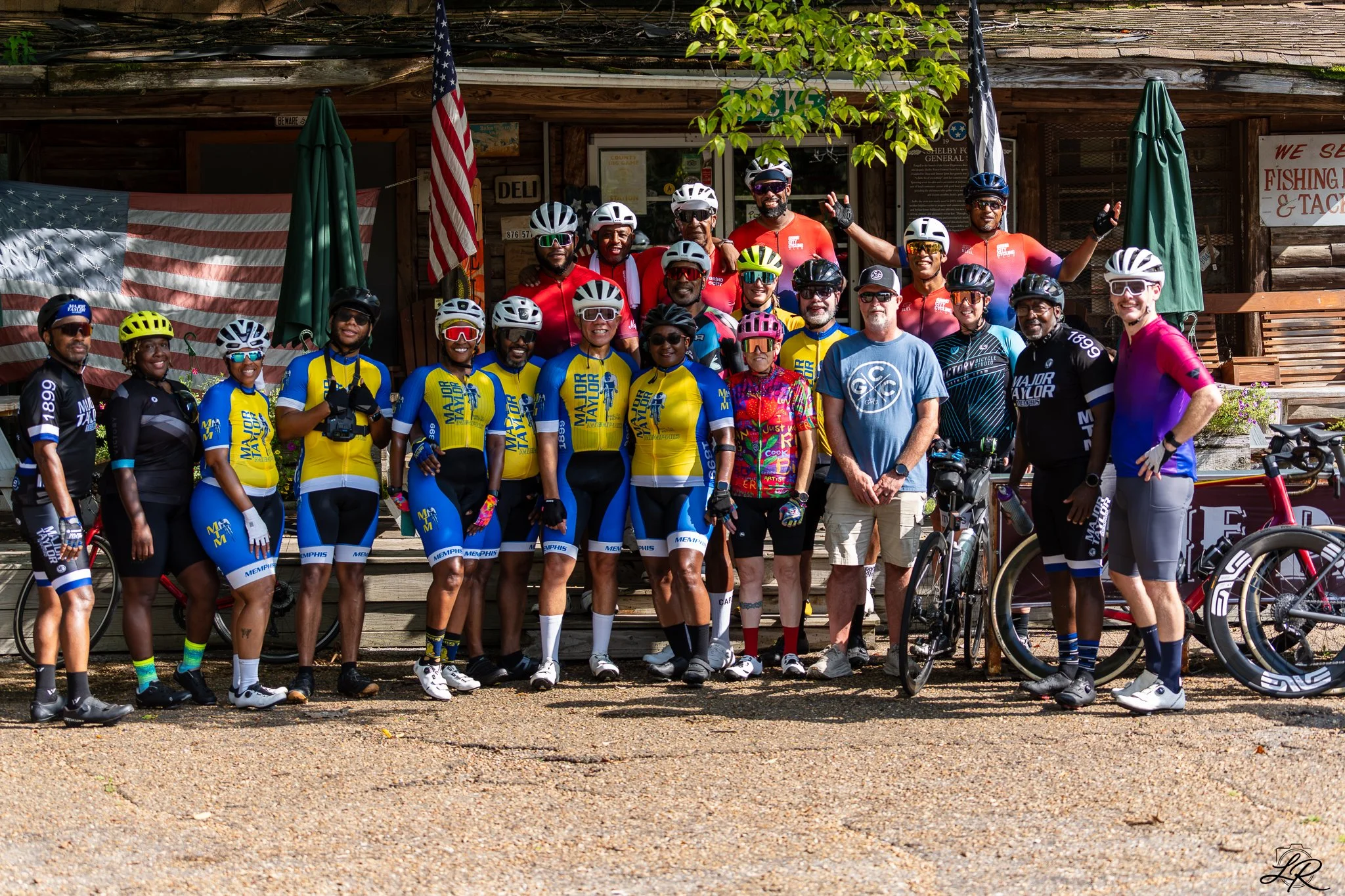 Group of cyclists and supporters posing in front of a wooden building with American flags, trees, and signs, smiling and waving at the camera.