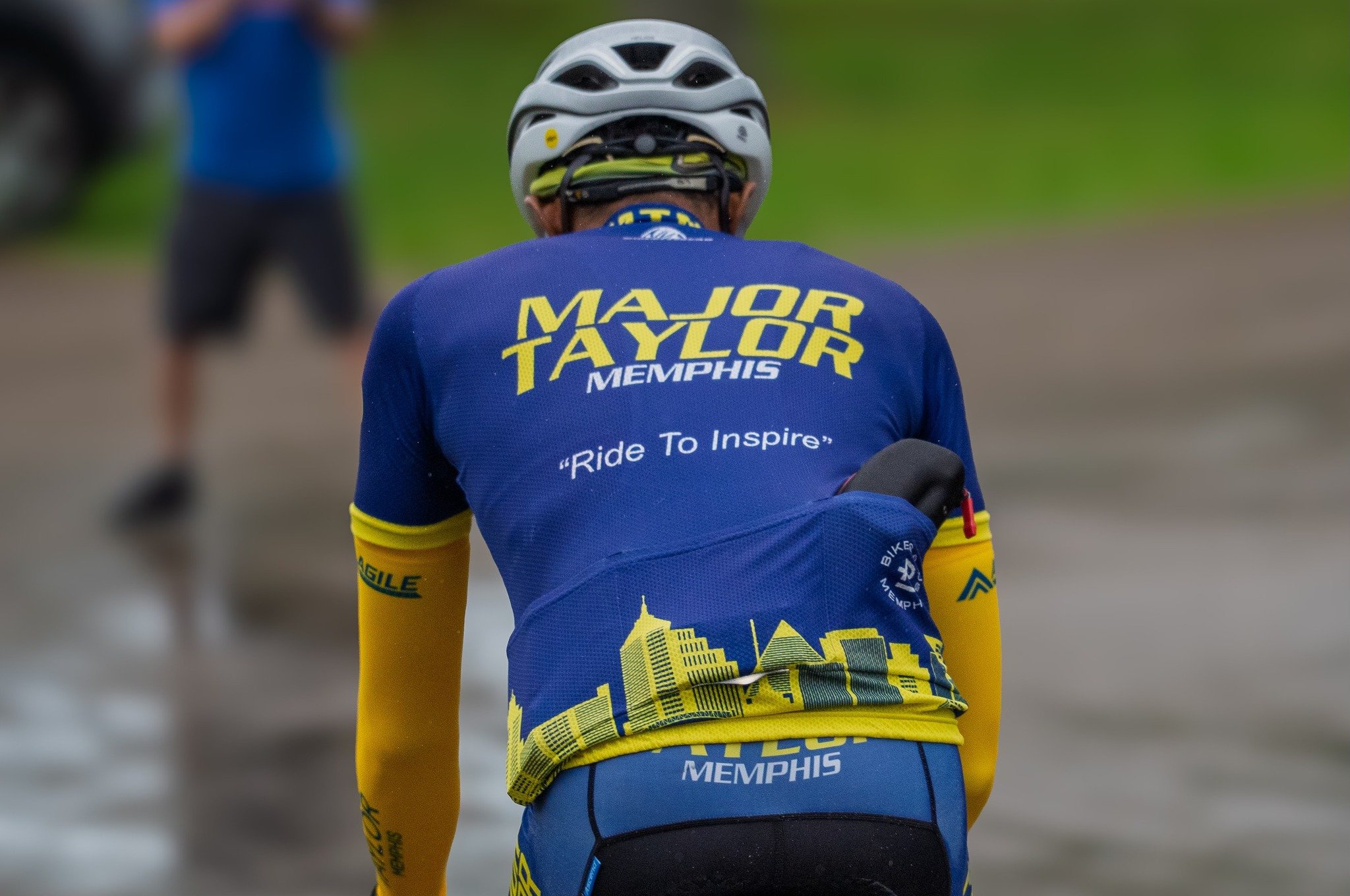 Back view of a cyclist wearing a blue and yellow Memphis Major Taylor cycling jersey and a white helmet, riding on a road with a blurred background.